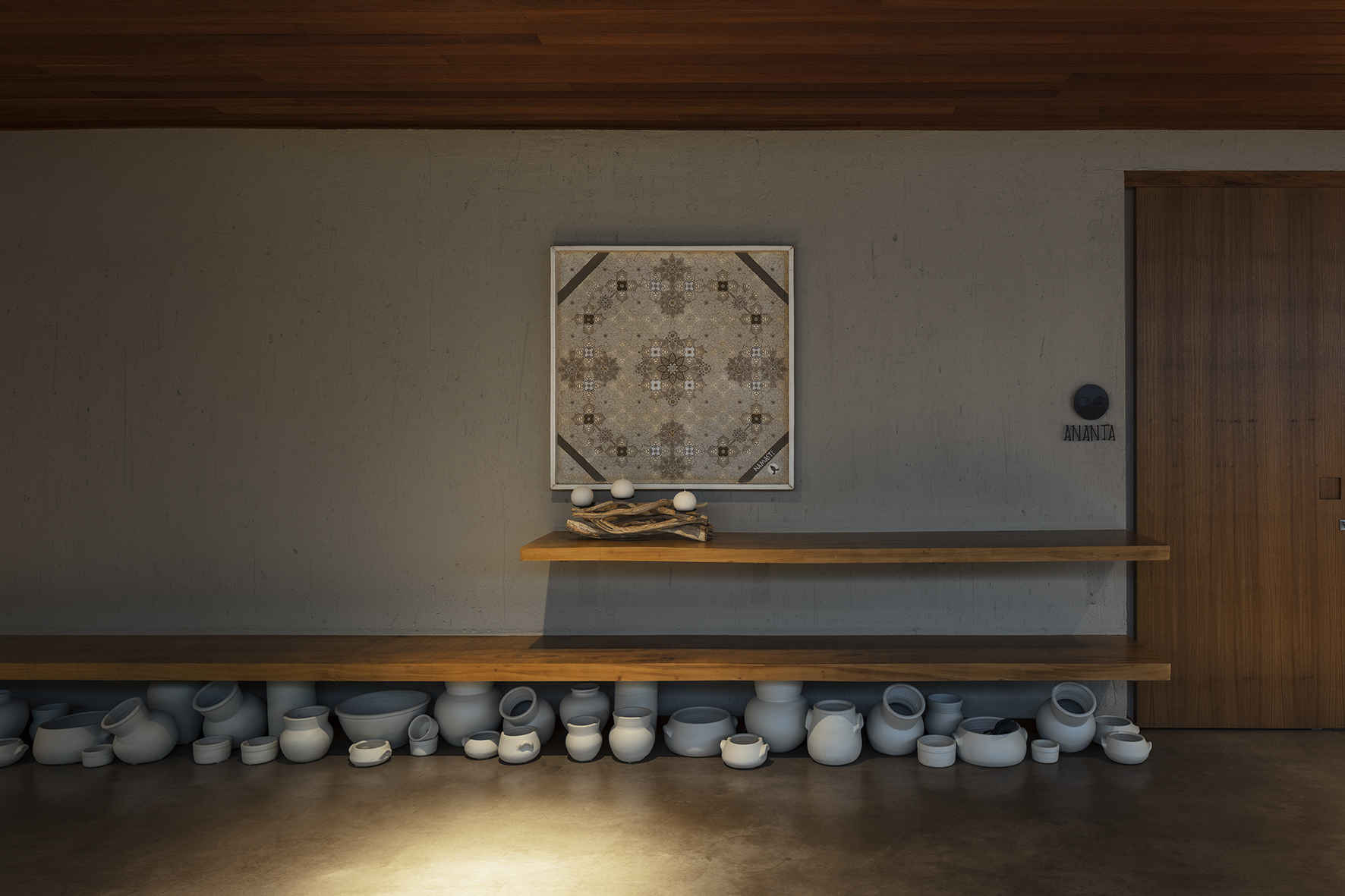 Wooden shelf above a row of white ceramic pots and bowls on the floor, a framed pattern hanging on a concrete wall