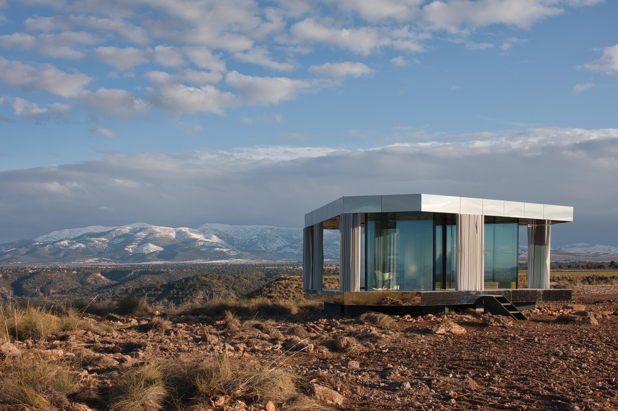 Mirrored glass pavilion with a flat metal roof and pale curtains on rocky ground, snowy mountains behind under broken cloud