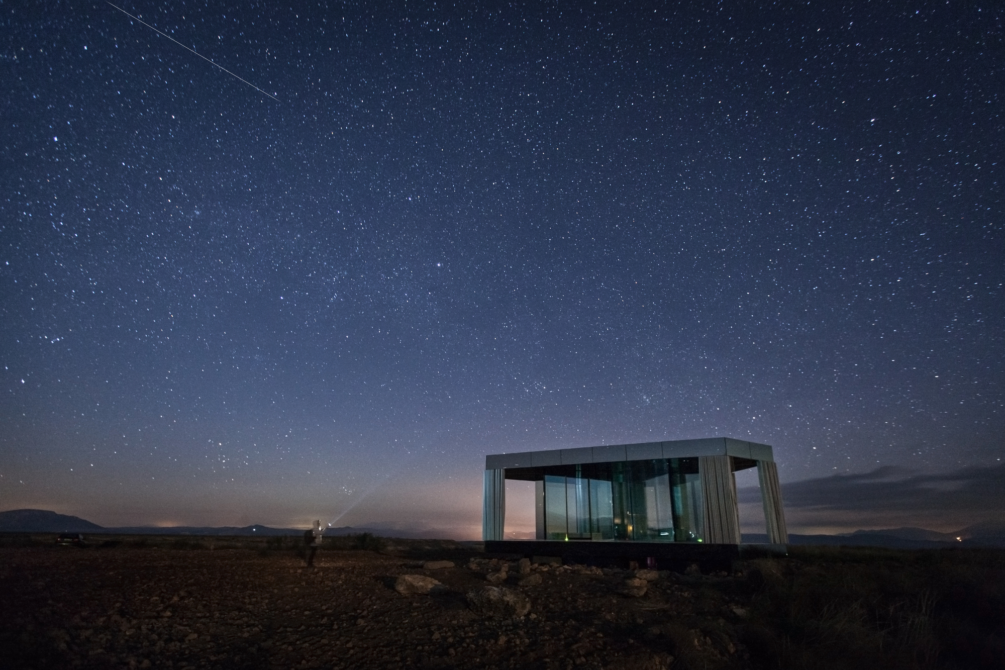 Glass pavilion glowing faintly beneath a dense starry sky, with a lone figure holding a light on the dark plain