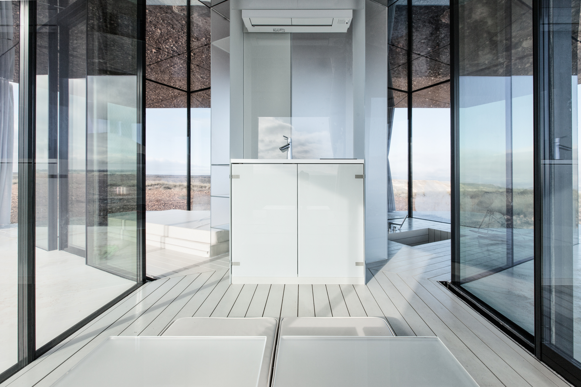 White vanity cabinet with a tap at the centre of the glass room, reflective ceiling and pale deck boards around it