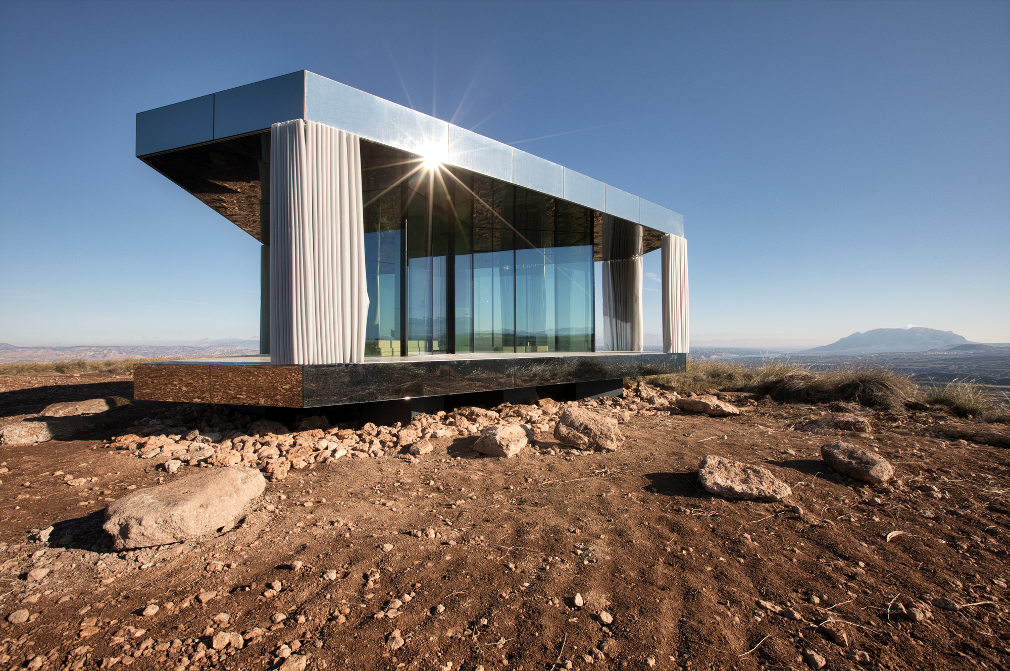 Low angle of the mirrored pavilion with draped curtains at the corners and the sun bursting over its roof edge