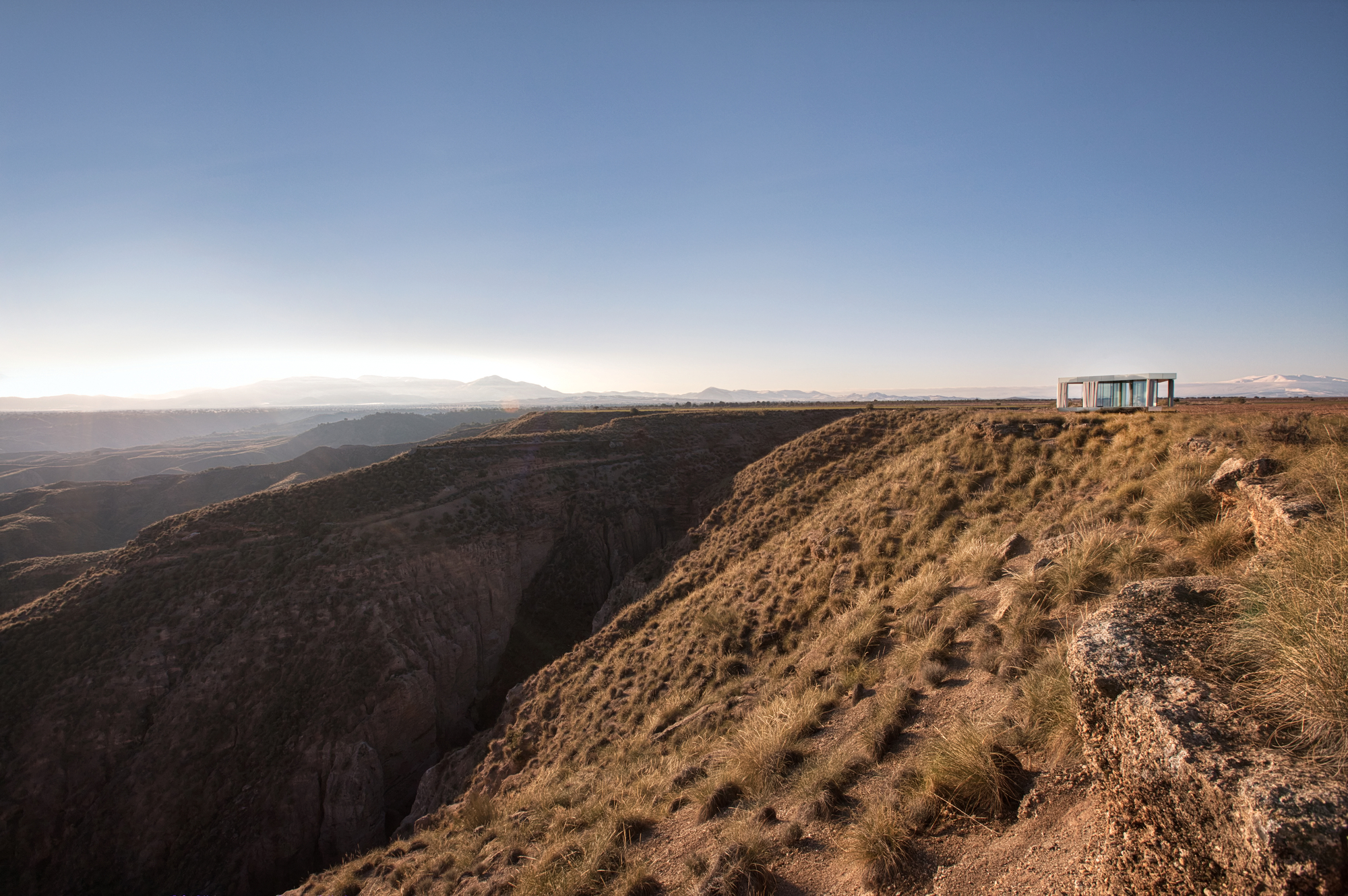 Distant glass pavilion perched on a canyon rim above eroded grassy slopes in low evening light