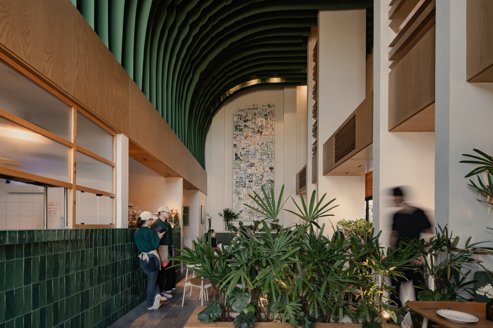 Restaurant aisle with green glazed tile counter, planted bed and a tall illustrated mural beneath a curved green ribbed ceiling, staff and a blurred passerby