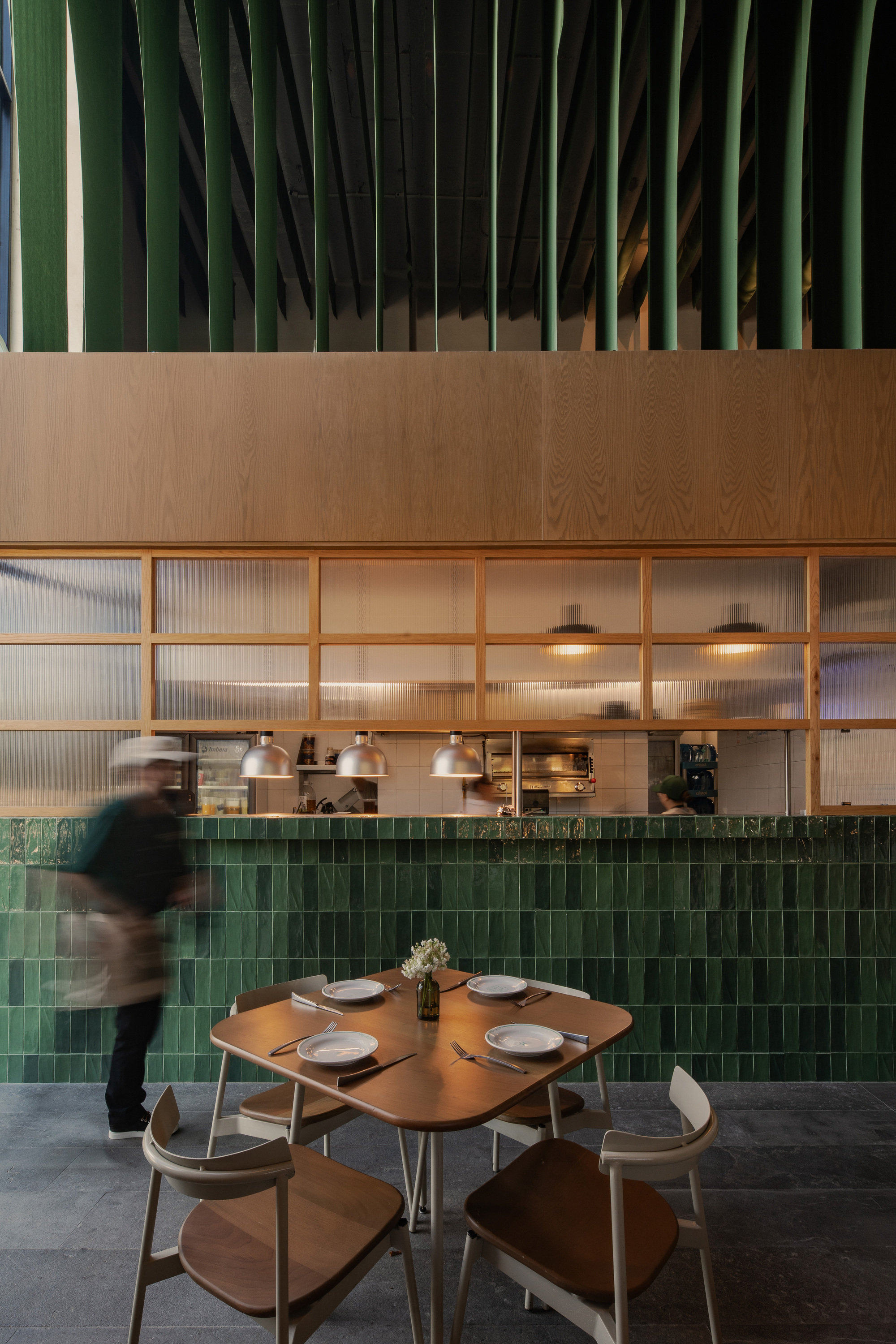 Wooden dining table and chairs facing an open kitchen with green tile counter, pendant lamps and a blurred server passing