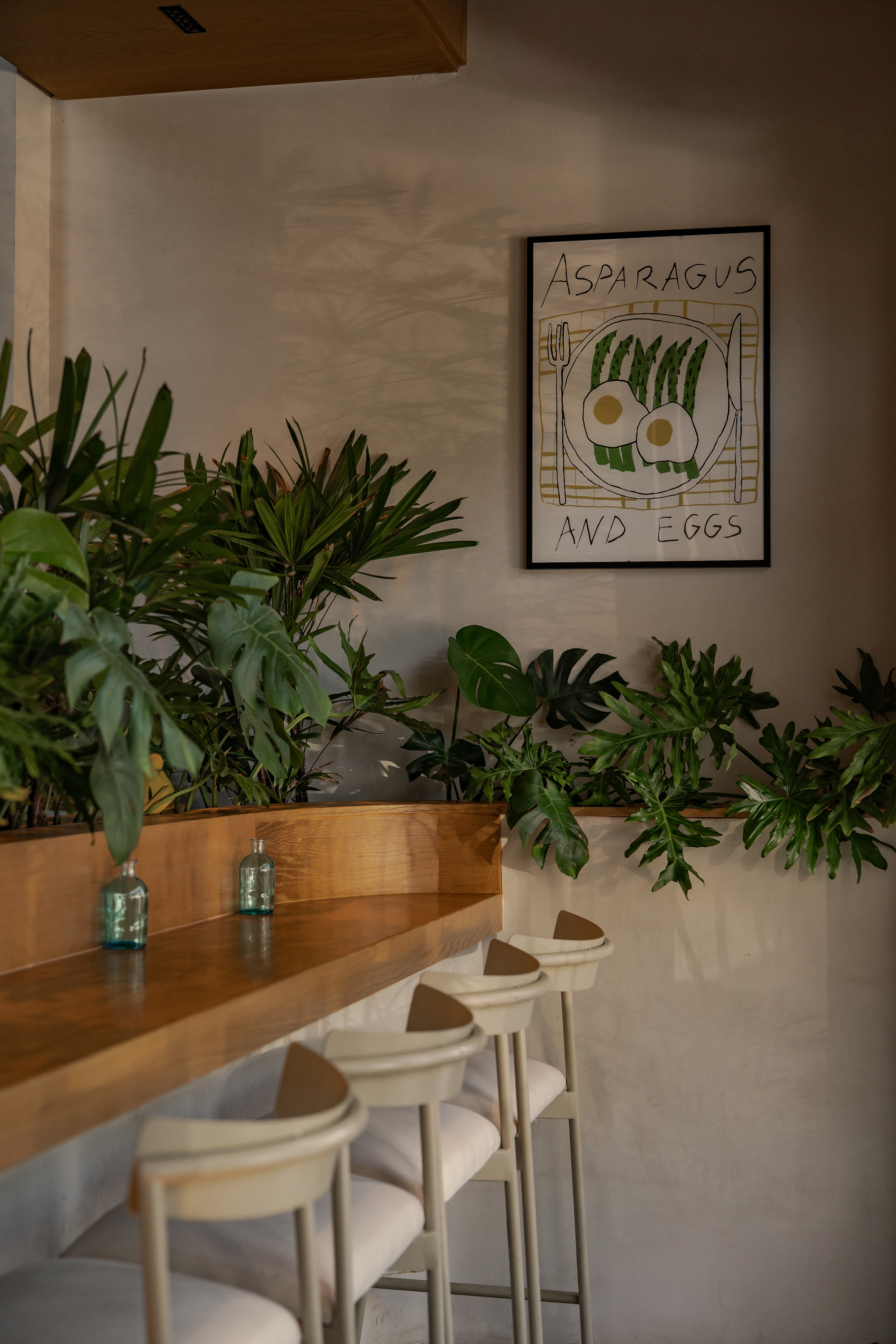 Timber bar counter with cream stools, potted foliage and a framed asparagus and eggs print on a plaster wall in soft light