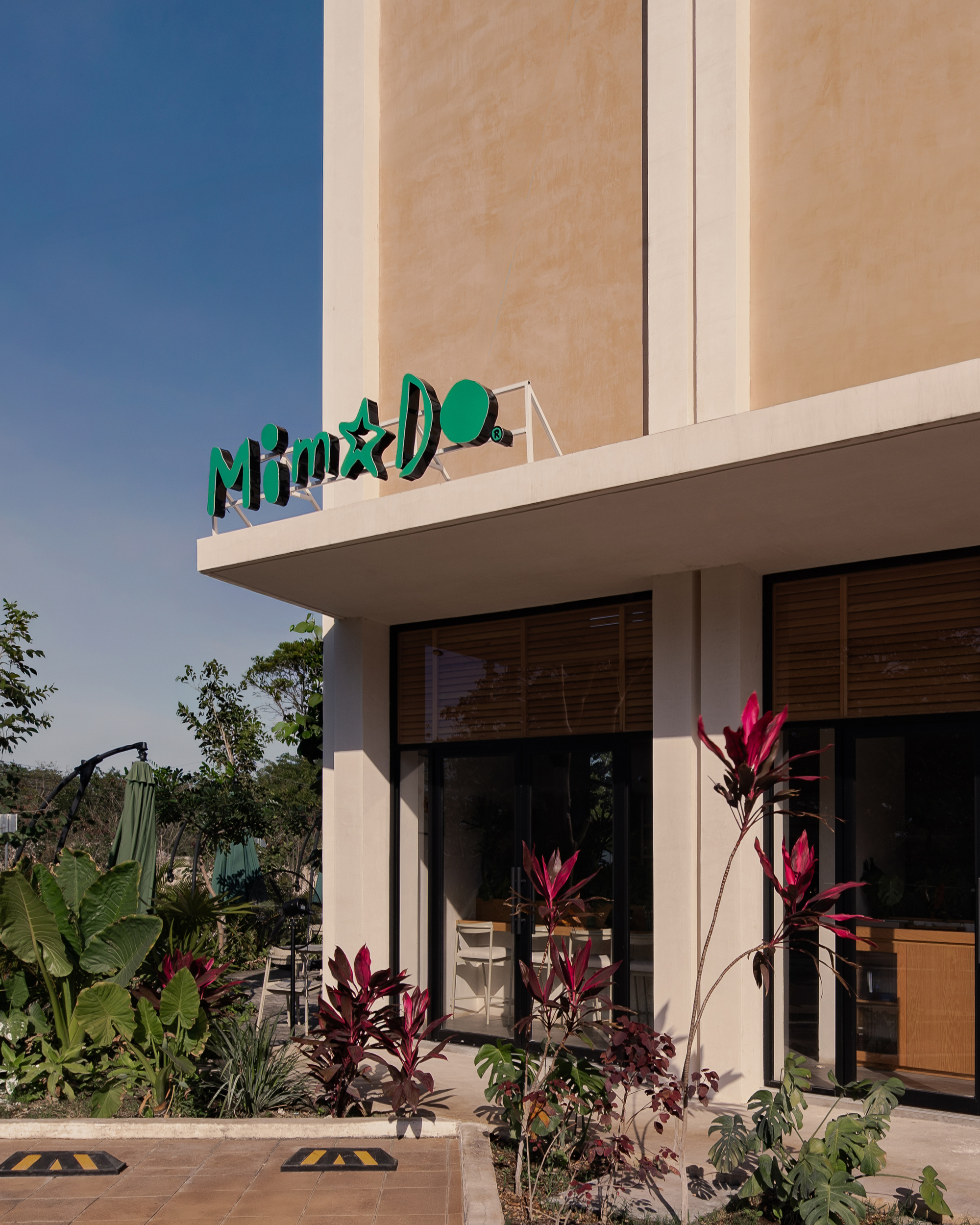 Corner of the two-storey facade with green rooftop lettering, cantilevered canopy and glazed storefront behind red-leaved plants under a clear blue sky