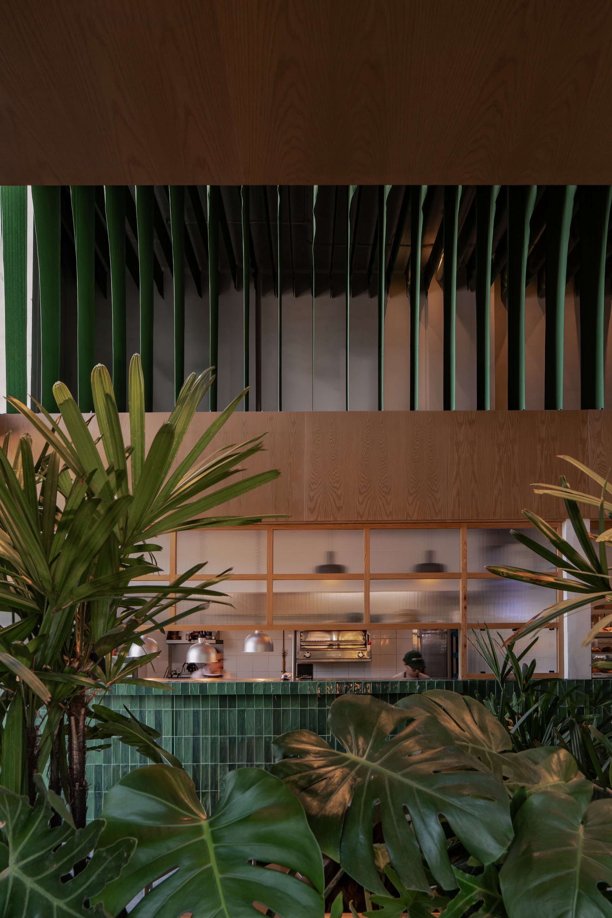 Open kitchen with green tile counter and timber frame glimpsed through monstera and palm leaves, with green ceiling fins above