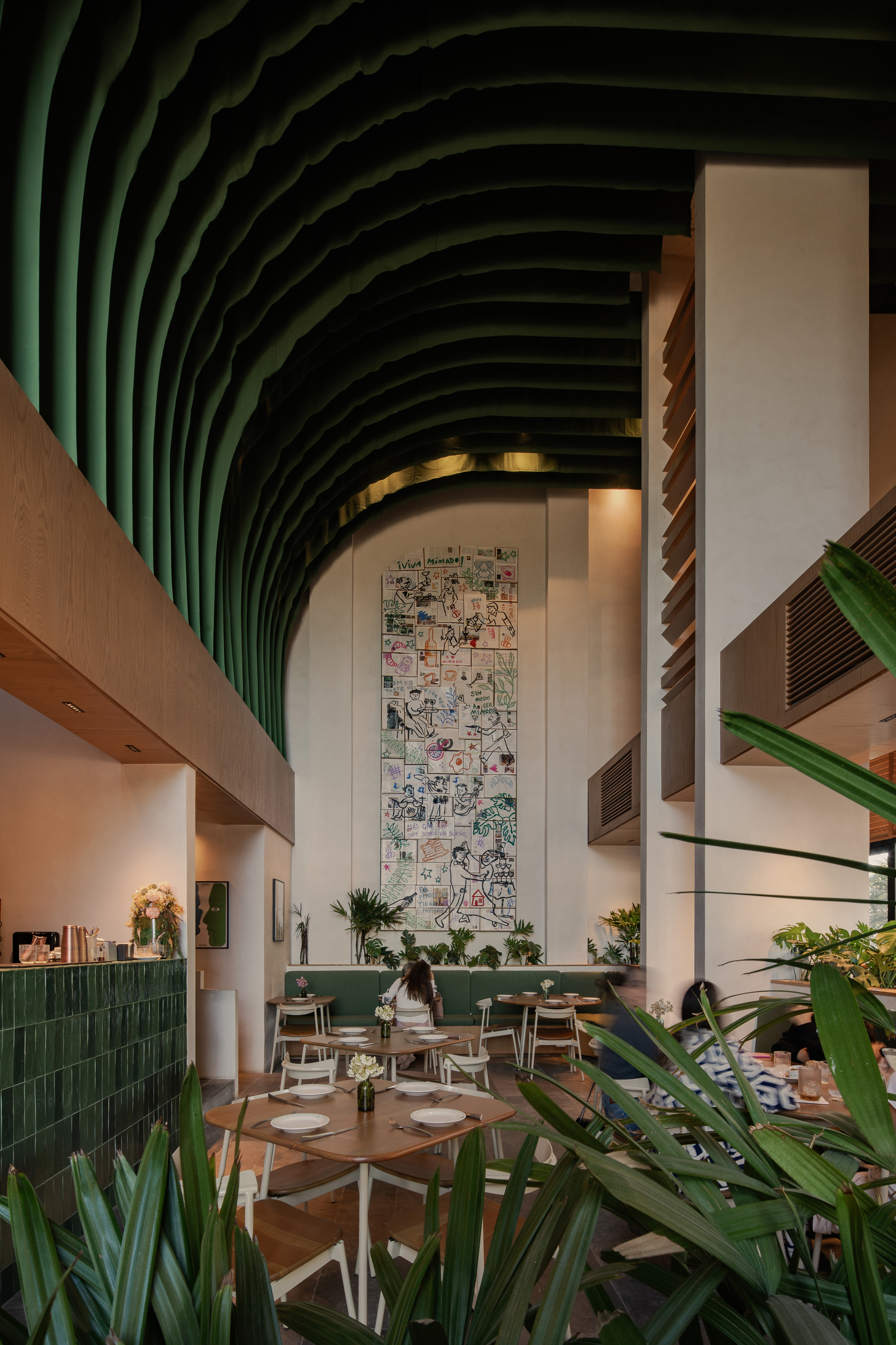 Tall dining hall with arched green ribbed ceiling, mural wall and set tables, palm fronds in the foreground in warm evening light