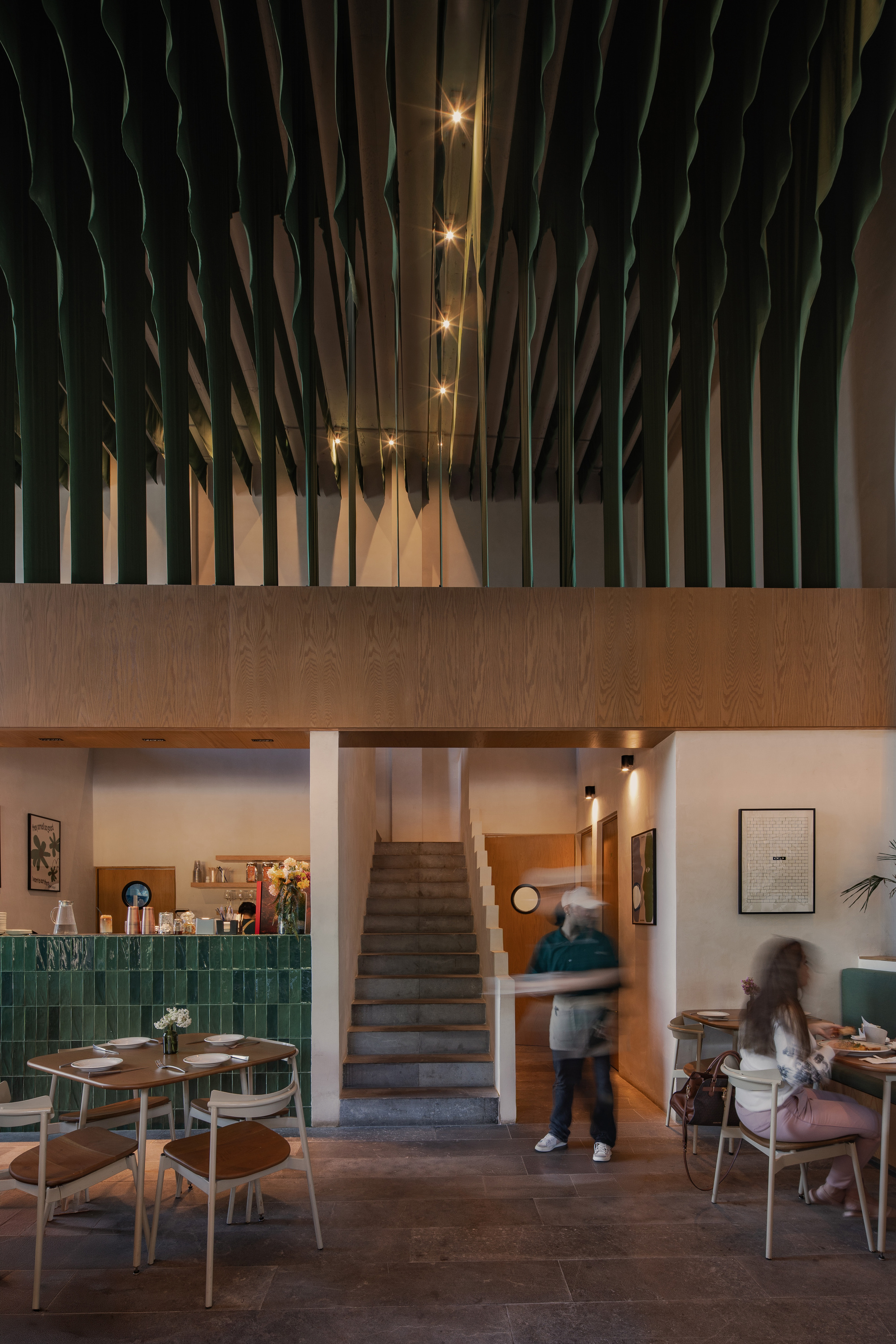 Stone stair rising between a green tiled counter and wood-clad mezzanine, with a blurred server and a seated diner under pendant lights