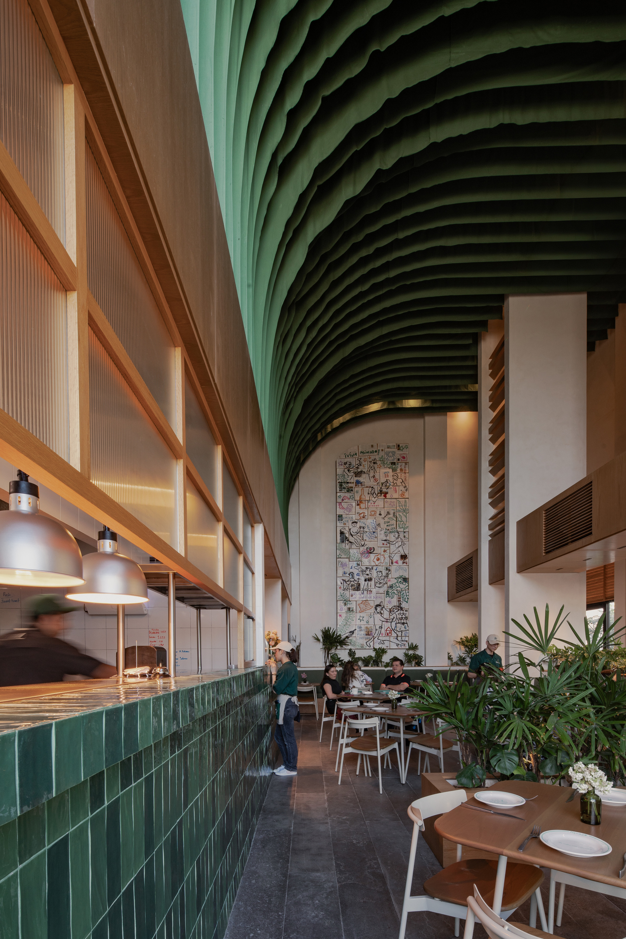 Long dining hall with a green tiled counter, pendant lamps and a vaulted fabric ceiling above a tall mural wall