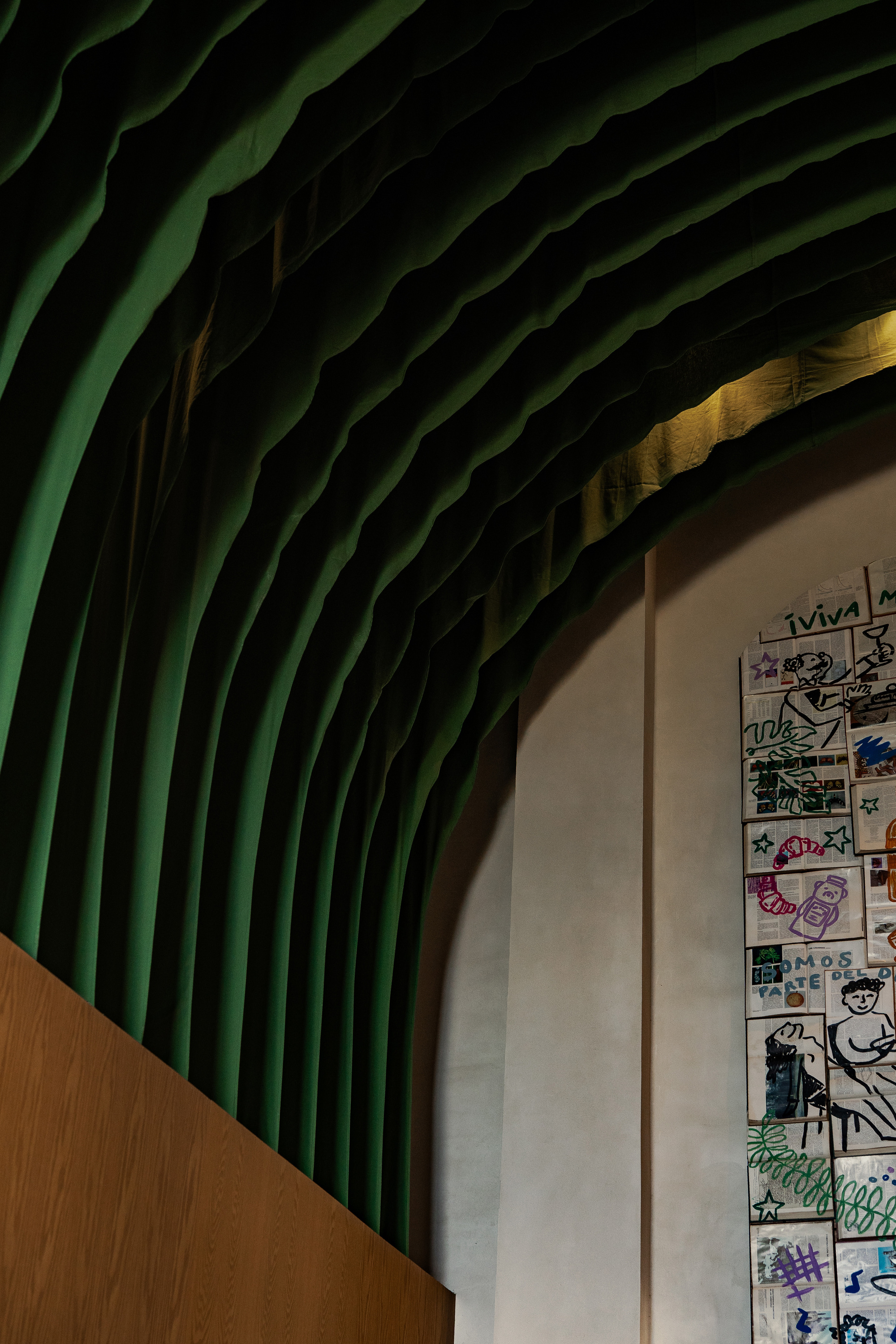 Close-up of the green pleated fabric vault meeting a plaster wall hung with small illustrated panels, a warm oak edge in the corner