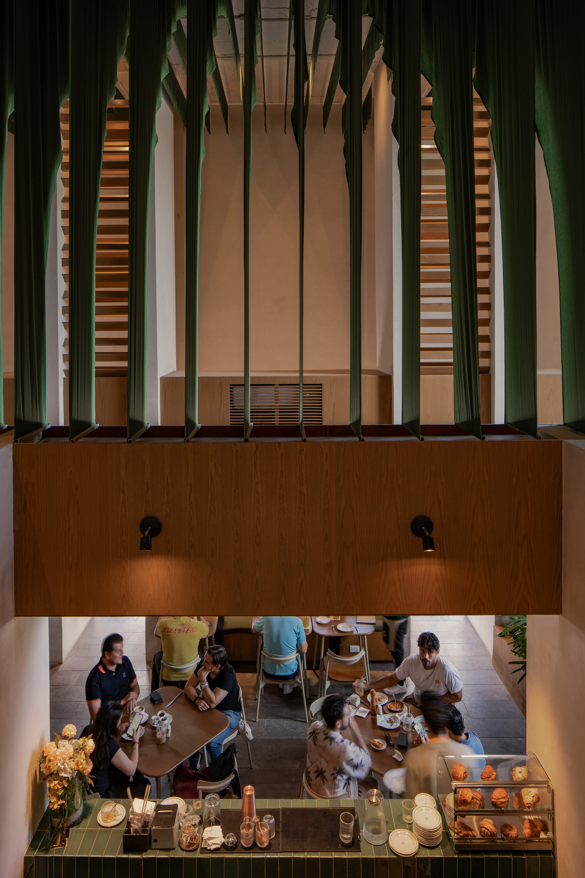 Looking down from above a timber mezzanine band at diners seated near a green tiled counter and a pastry display case