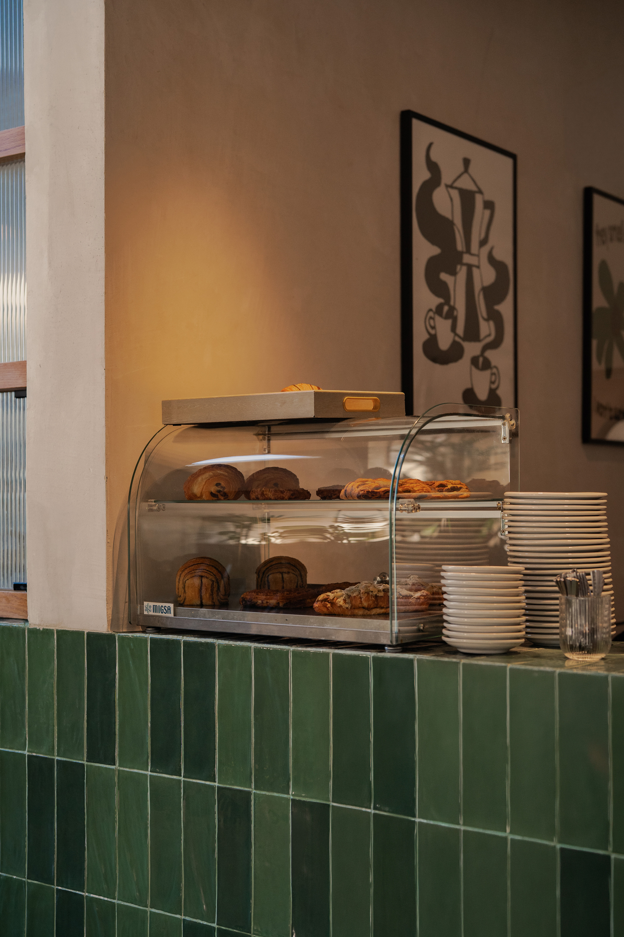 Glass pastry display case on a green glazed tile counter beside stacked plates and a framed coffee pot print in warm light
