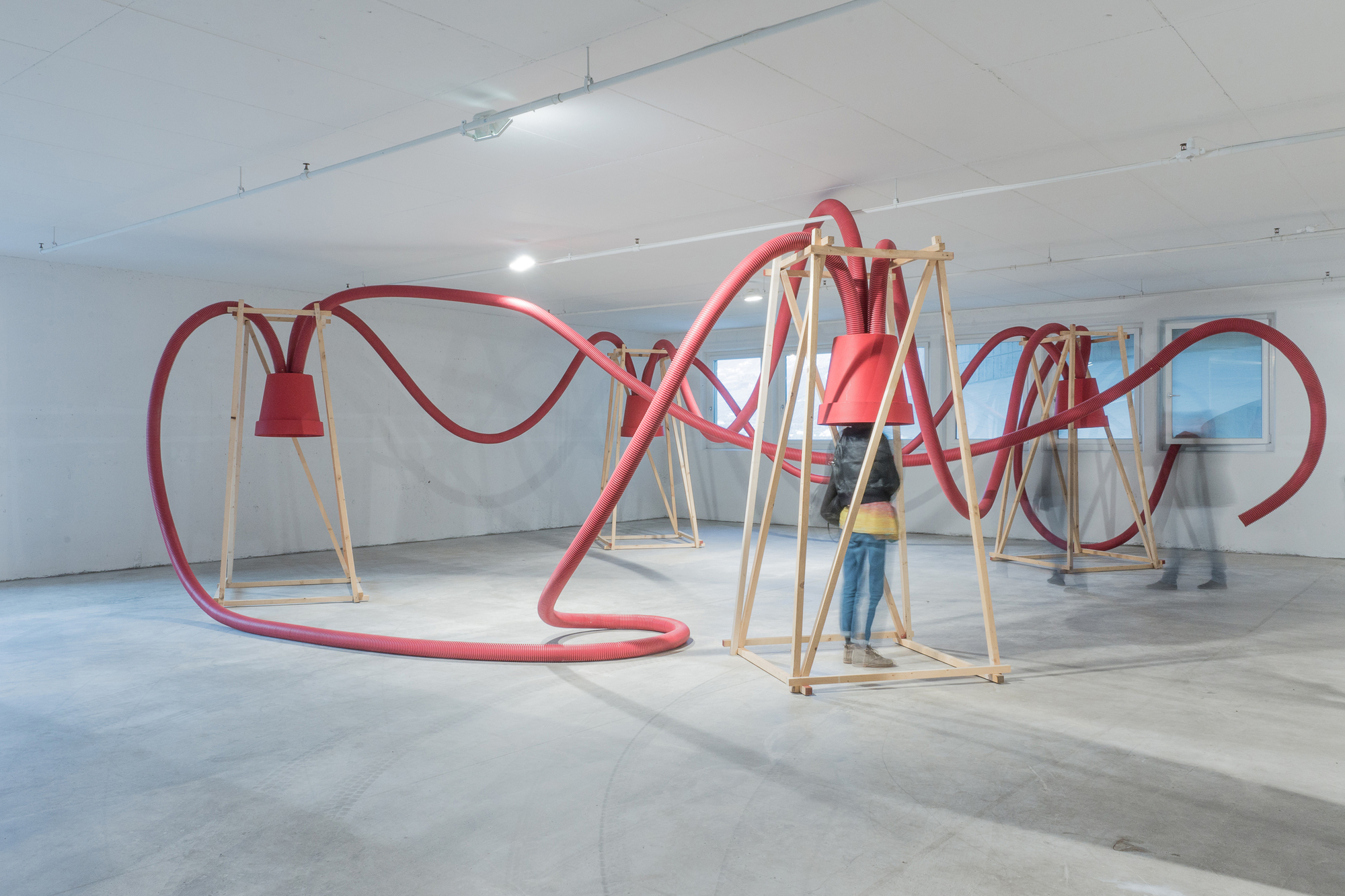 Red corrugated hoses looping between timber frames topped with red funnels, with a visitor standing inside one frame in a white room