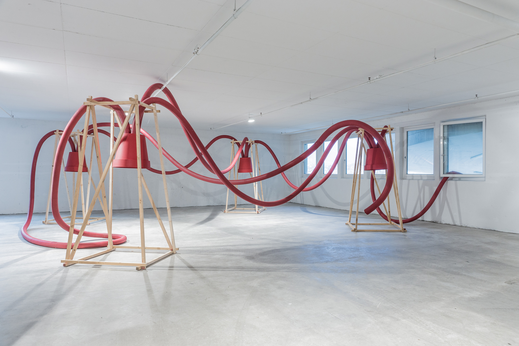 Wide view of red hoses draping across three timber frames in an empty room with a concrete floor and ribbon windows