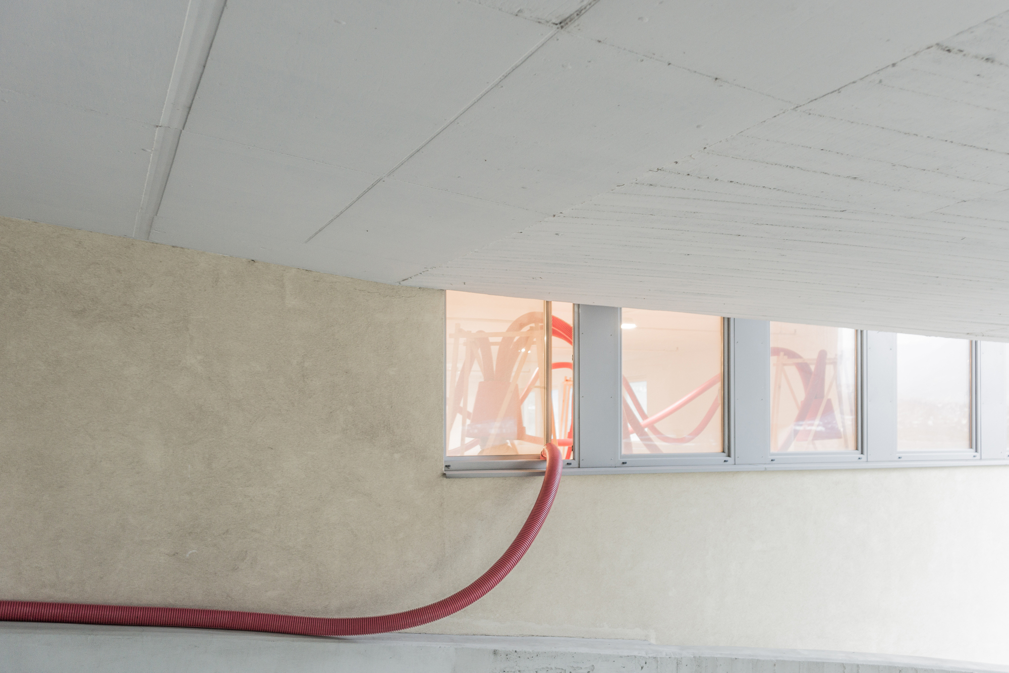 Red hose curving up a plaster wall to a ribbon window, with the installation reflected in the glass