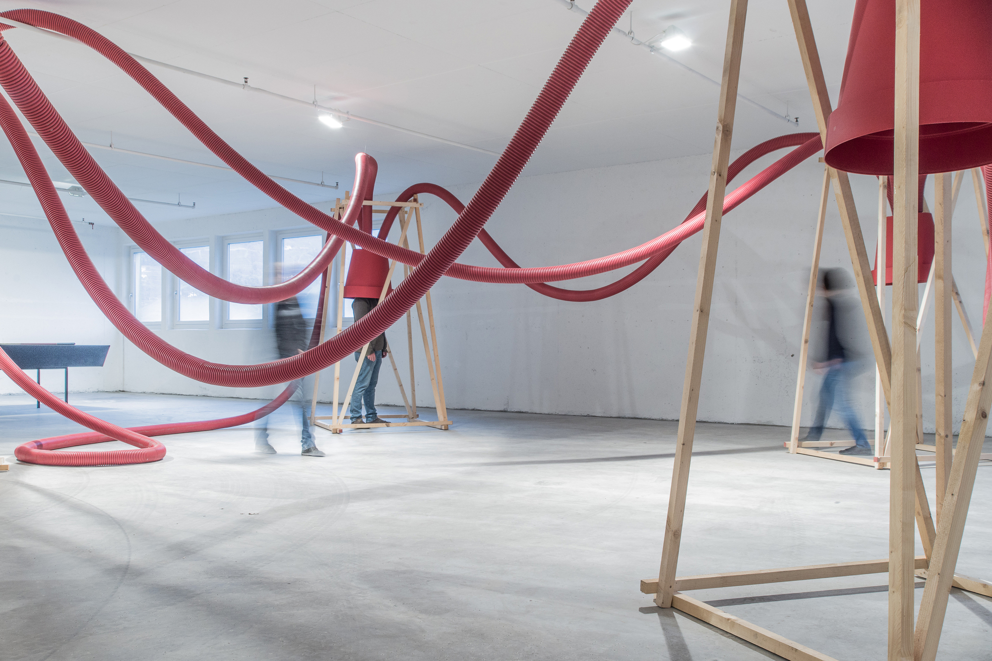 Low view past a timber frame and red funnel toward hoses sweeping across the room, with blurred visitors walking by