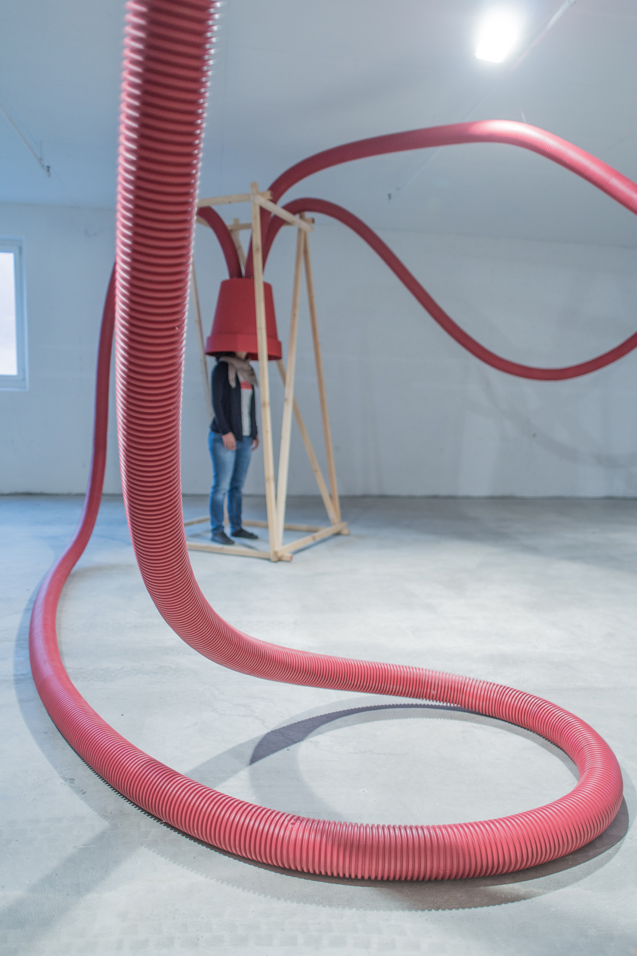 Looping red corrugated hose curling across a concrete floor toward a timber frame with a red bucket over a standing person's head