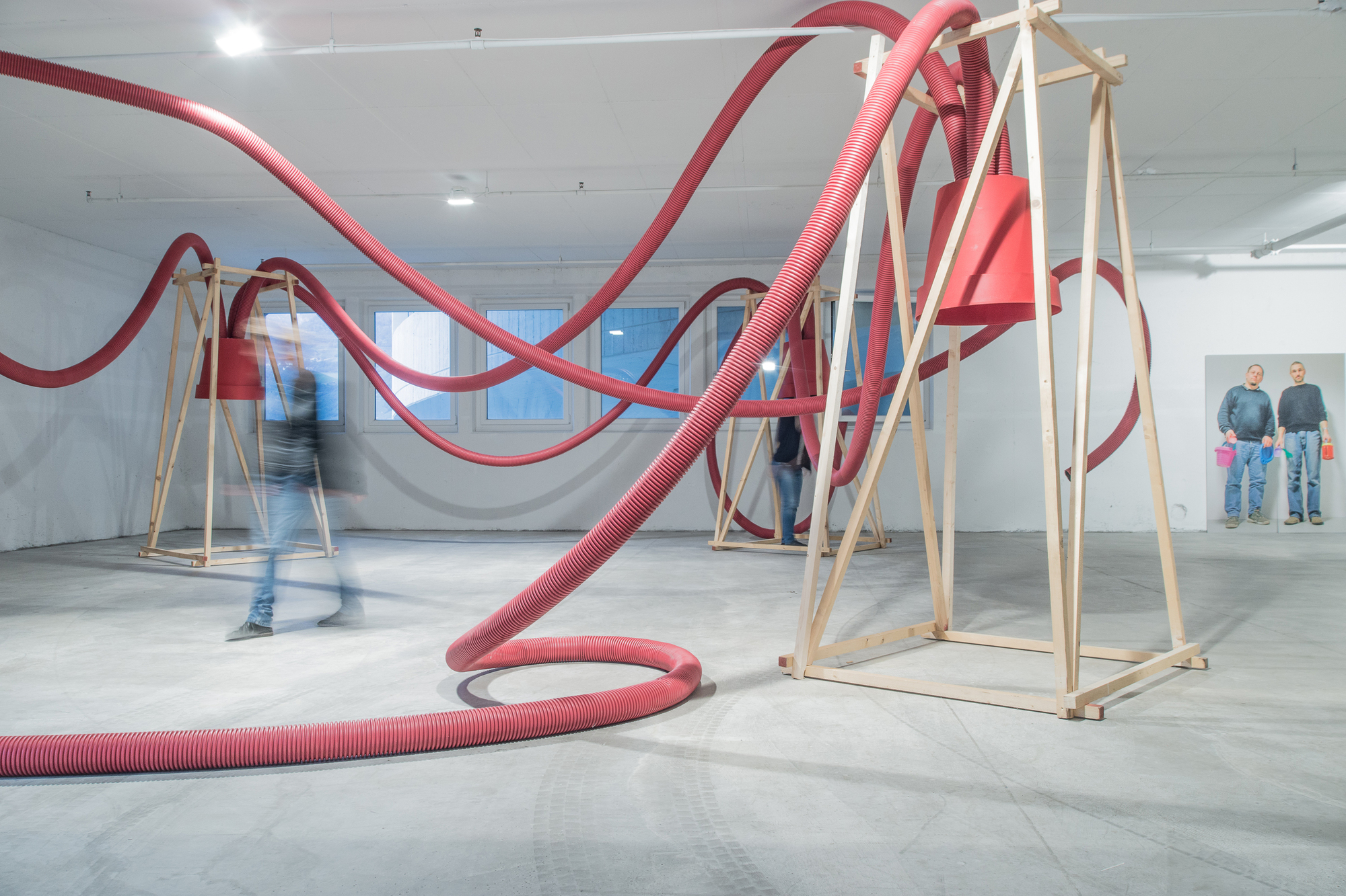 Red hoses sweeping between wooden A-frames in a white gallery, with a blurred walker crossing and a photo of two men on the wall