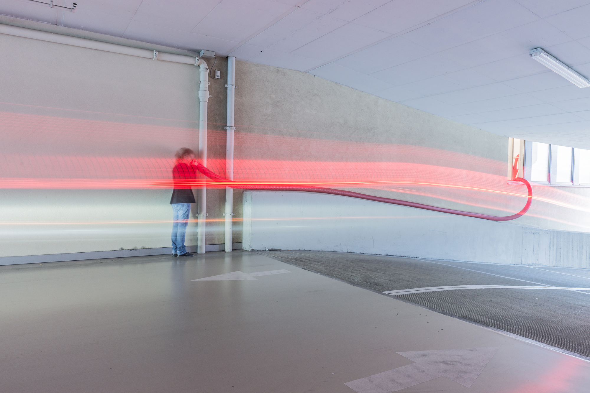 Long-exposure streak of red light trailing from a person holding a hose against a parking garage wall beside a ramp
