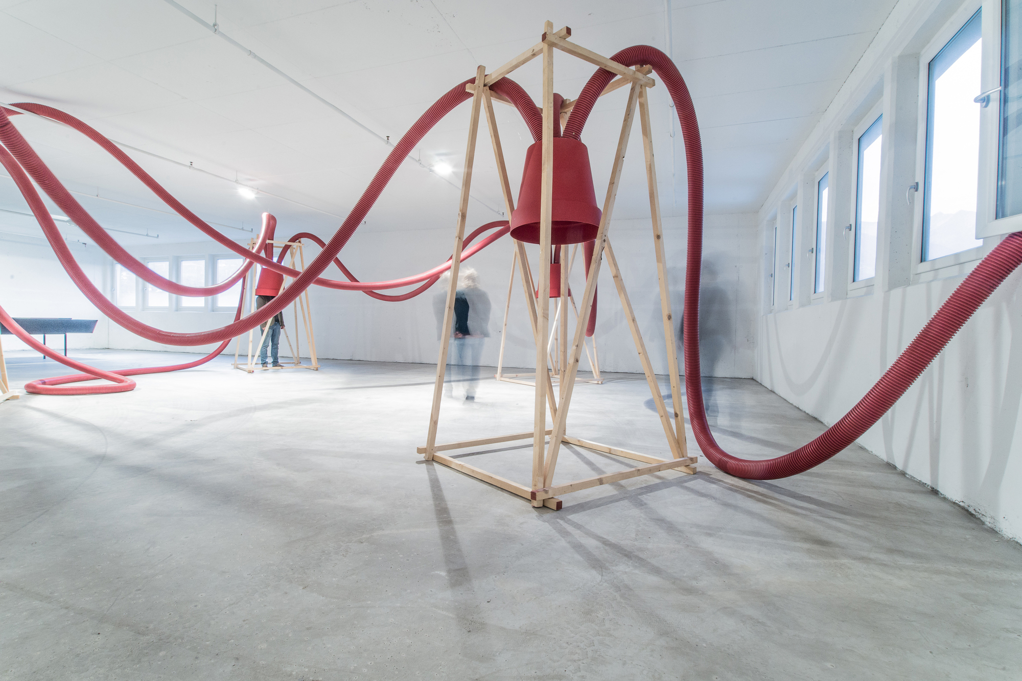 Wide view of a wooden frame holding a red bucket, with hoses arcing overhead past a row of windows and a blurred figure