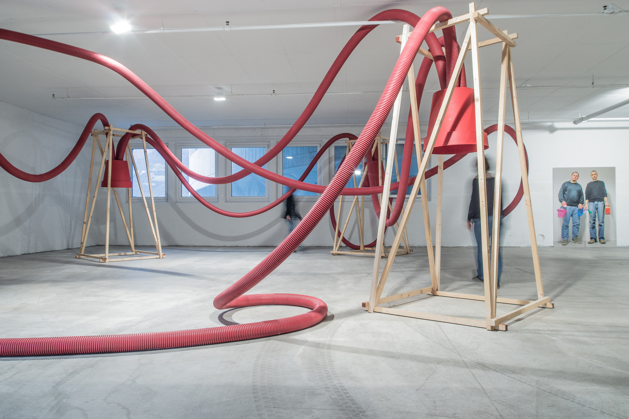 Red hoses looping over timber frames in a white room, with a blurred person inside one frame and a portrait of two men behind