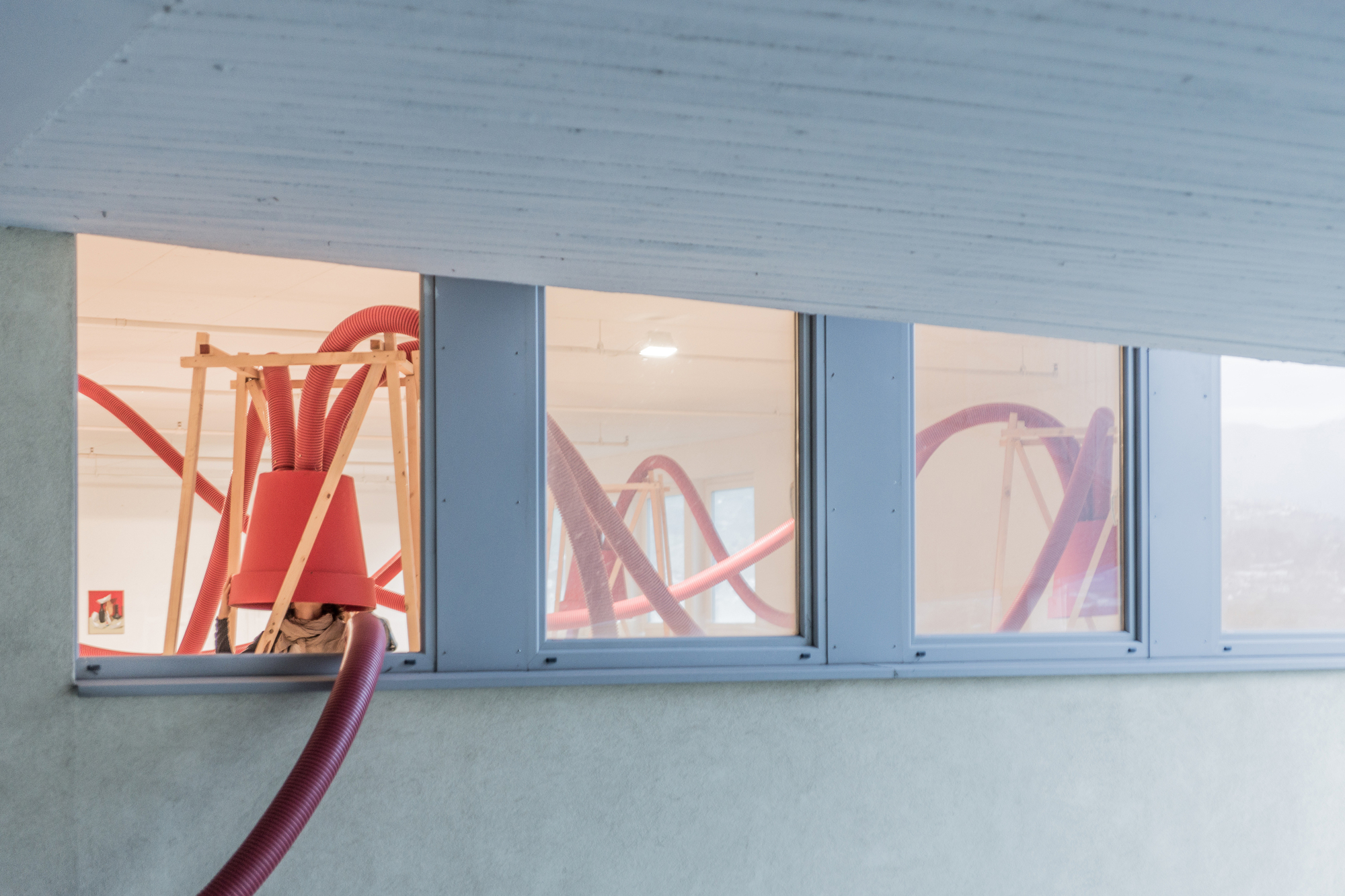 Red corrugated hoses looping through timber frames with inverted buckets, seen through a row of ribbed windows in soft daylight