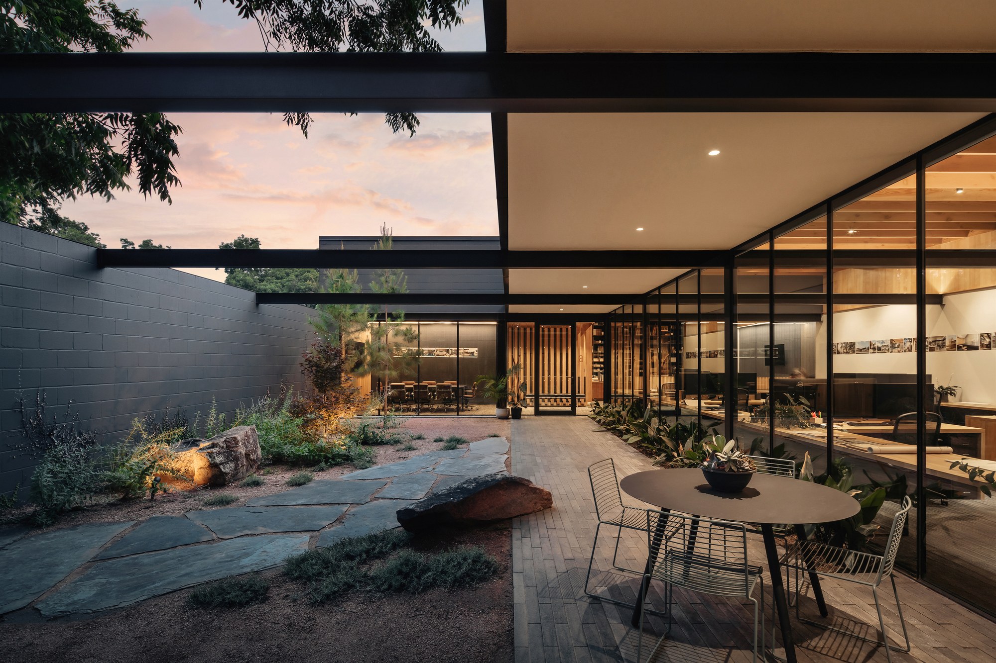 Courtyard with flagstone path, boulders and a round table beside a glazed office wall under a pink dusk sky