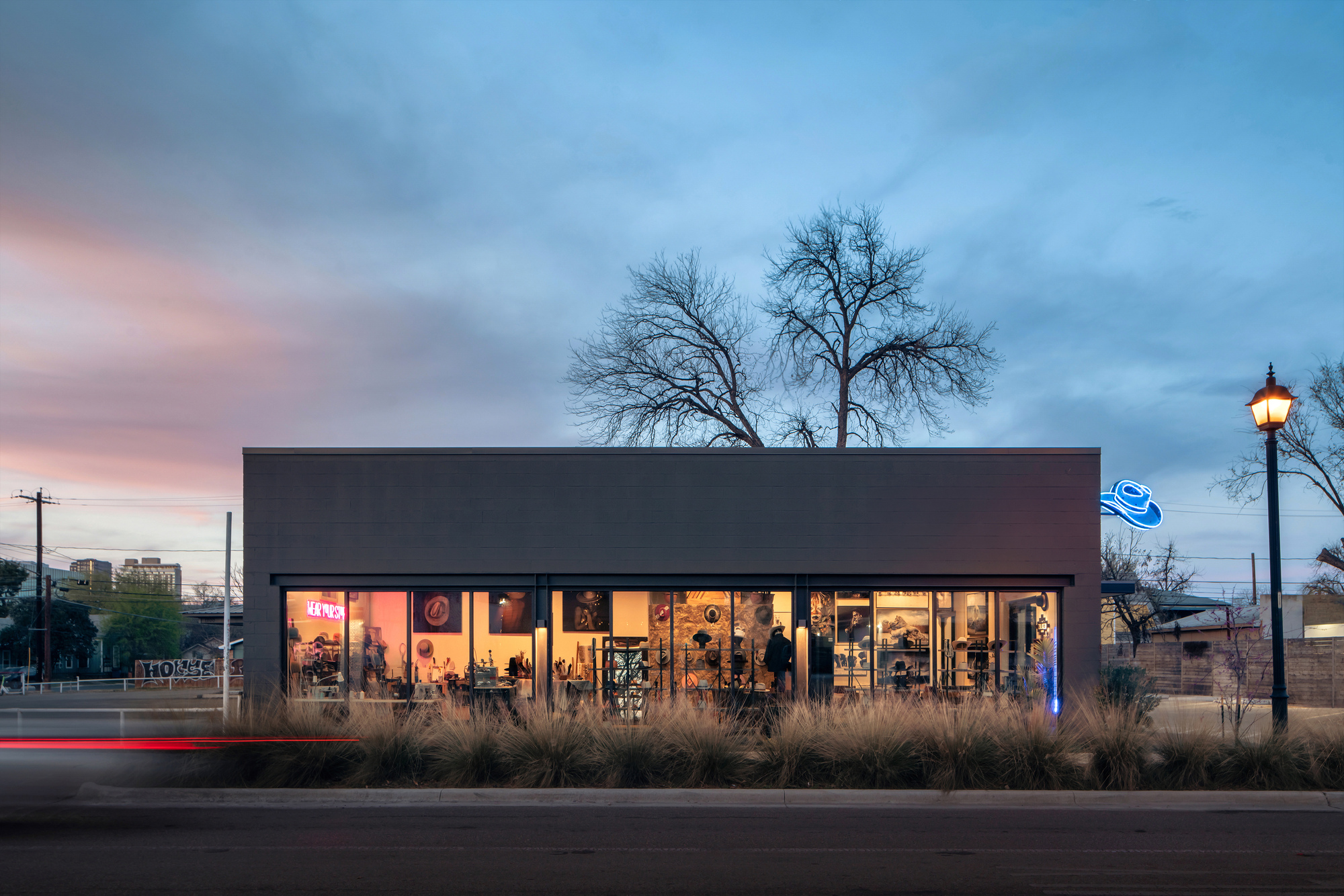 Street facade of a low dark box with glowing glazed storefront and a bare tree behind, seen at dusk