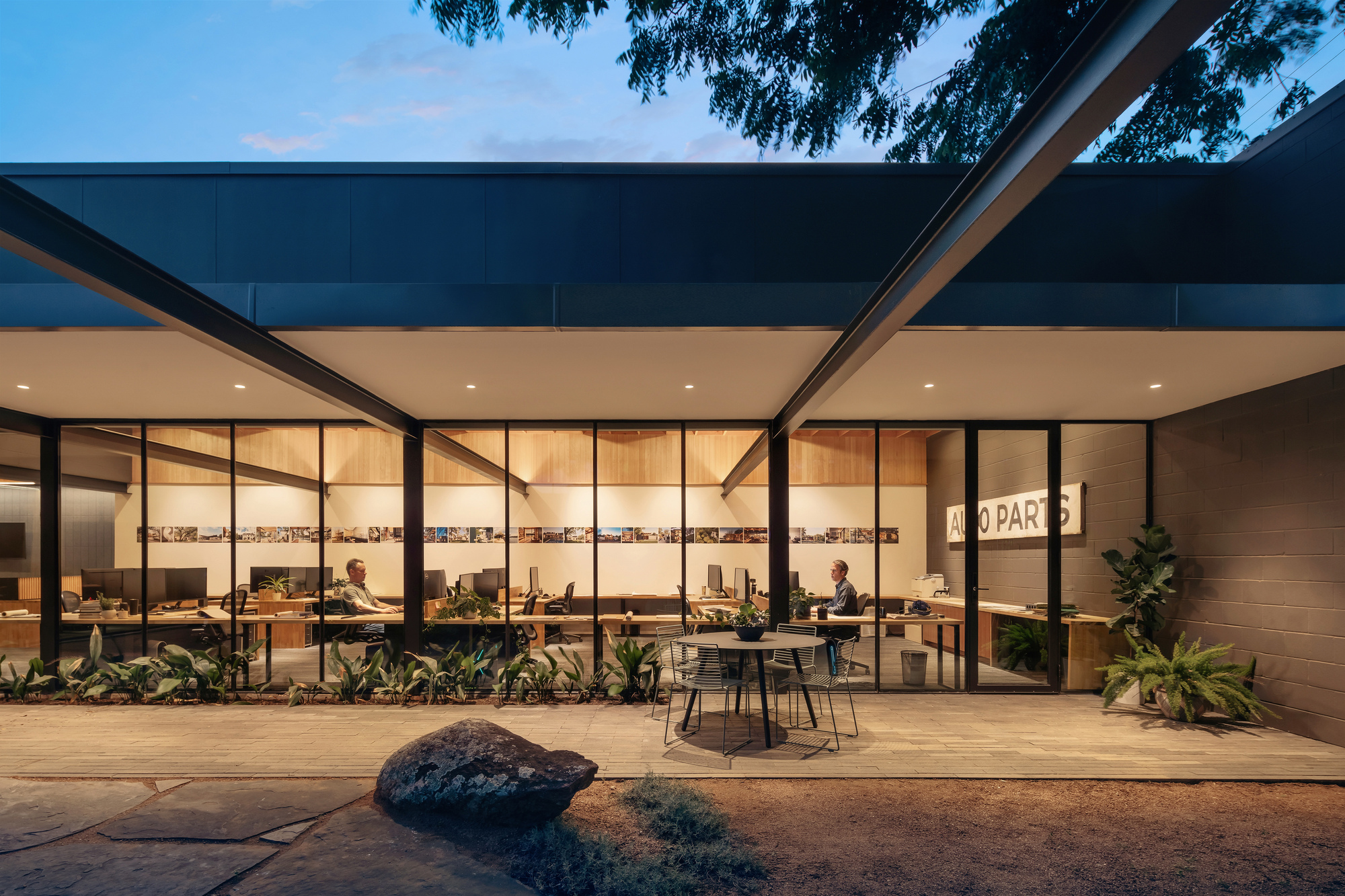 Glazed office wall opening onto a timber deck with a bcafé table and boulder, lit at twilight