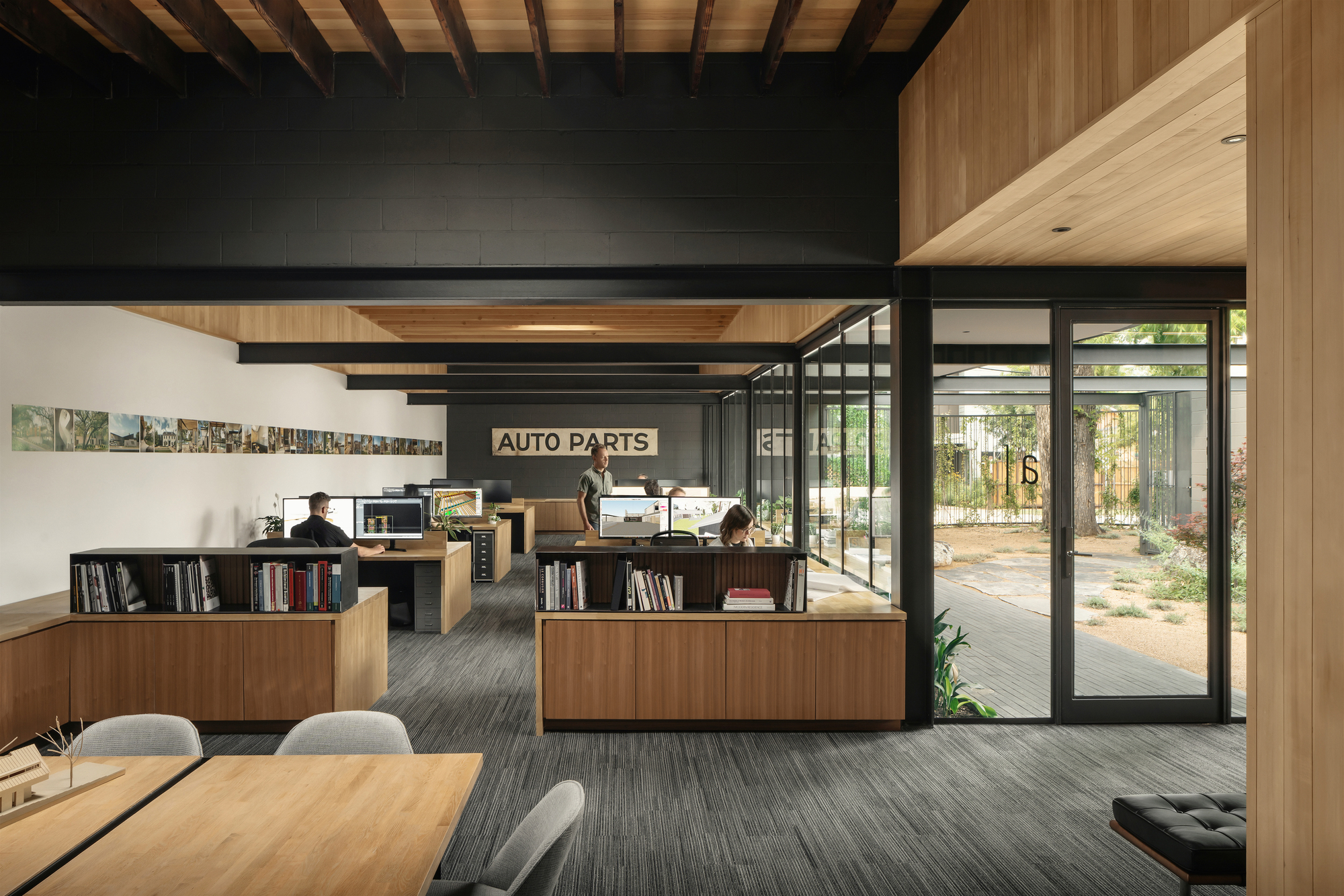 Open office interior with timber desks, low bookshelves and exposed rafters, glass doors opening to a sunlit courtyard