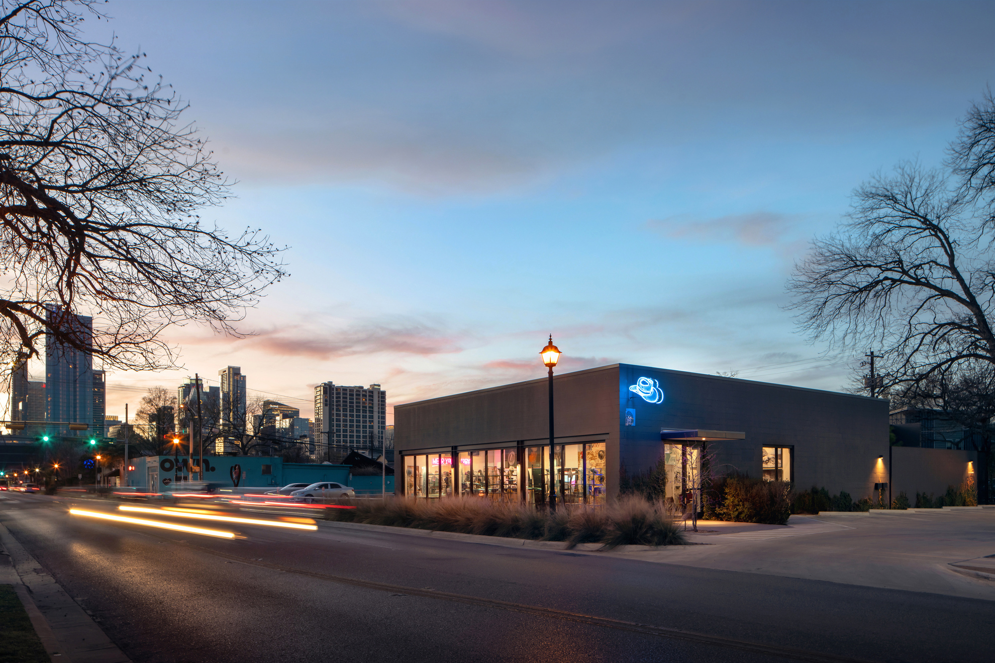 Corner view of the low gray building with blue neon hat sign, ornamental grasses and streaking car lights at dusk
