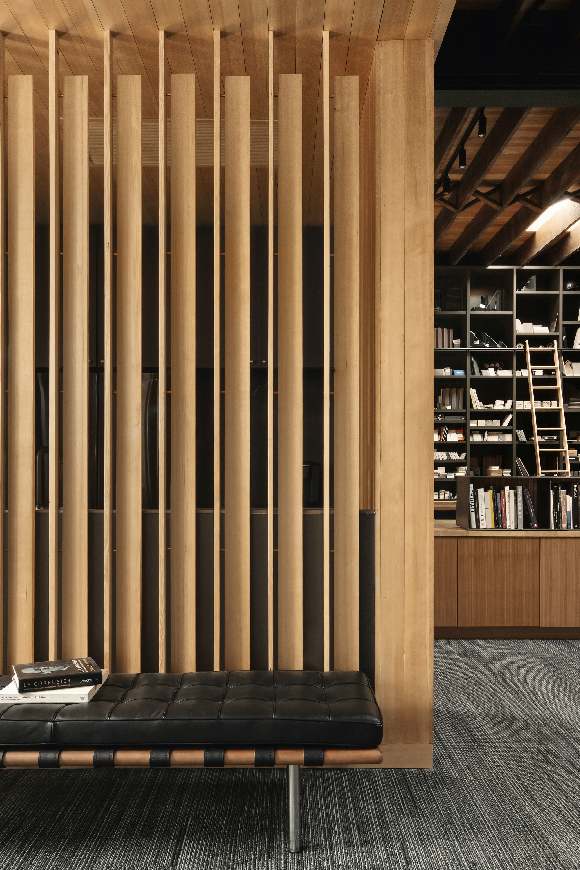 Vertical timber slat screen above a black leather bench with books, and a ladder shelf beyond in warm light