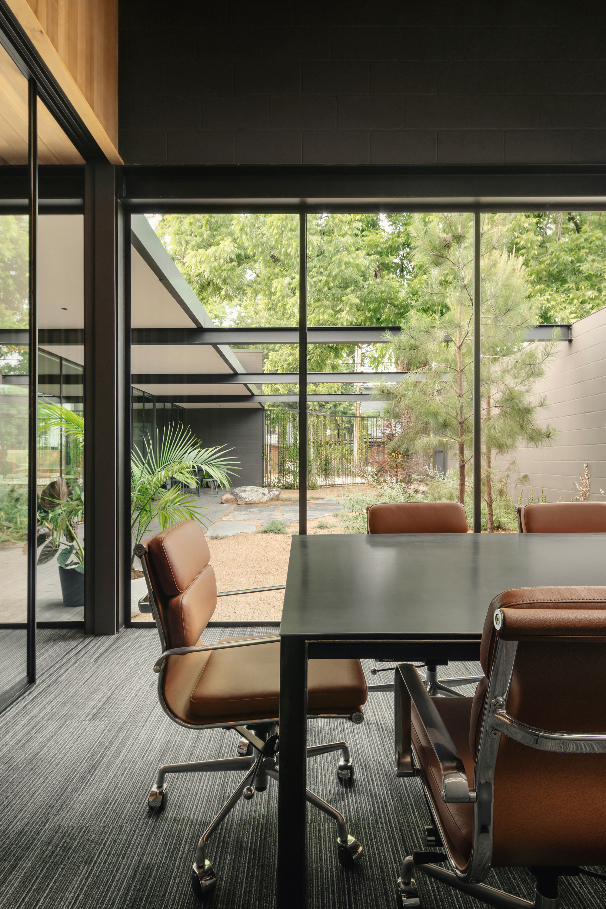 Conference table and tan leather chairs facing a glass wall onto a courtyard with pine trees in soft daylight