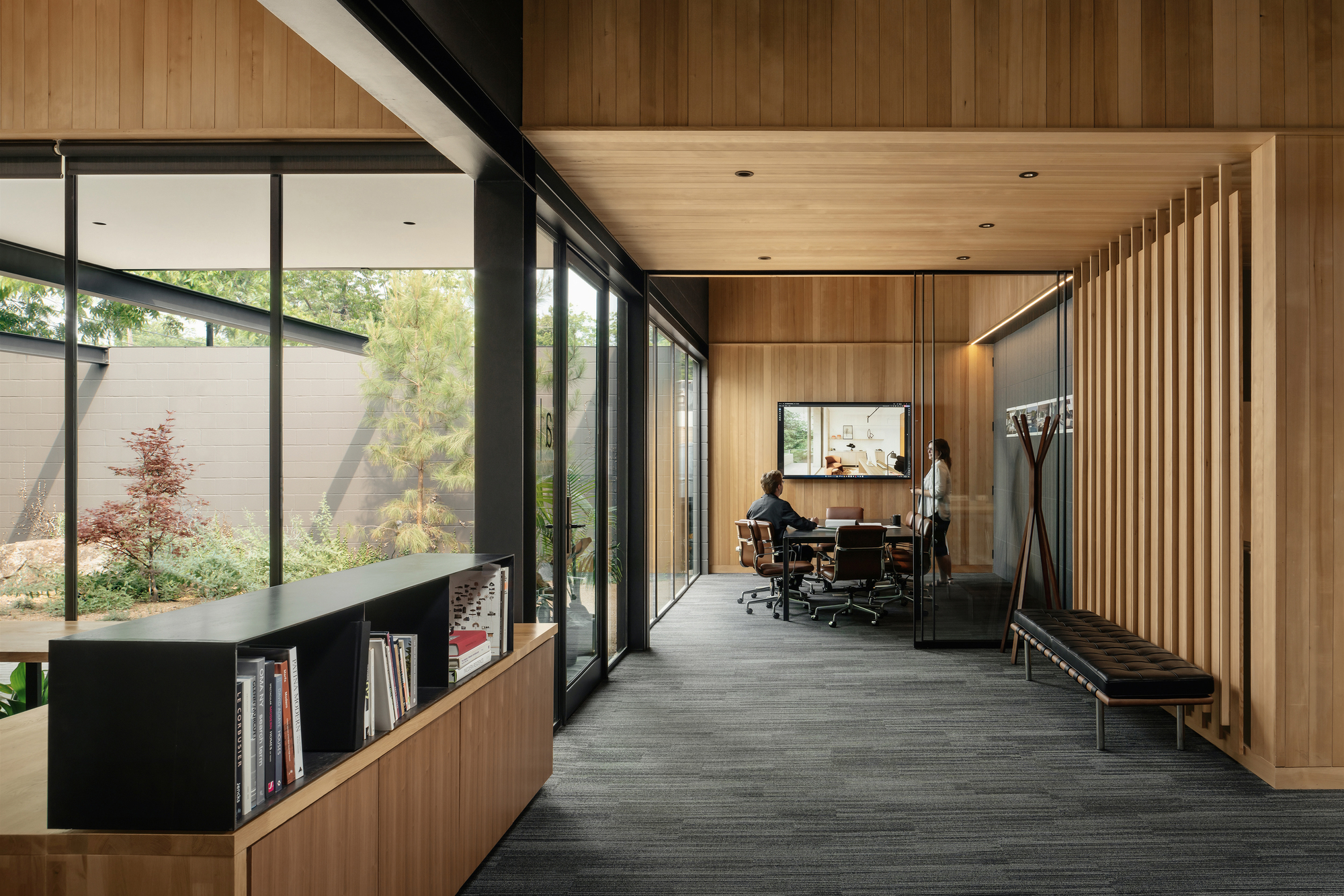 Timber-lined corridor with a built-in bookshelf, slatted wall and bench, leading to a glazed meeting room where two people talk