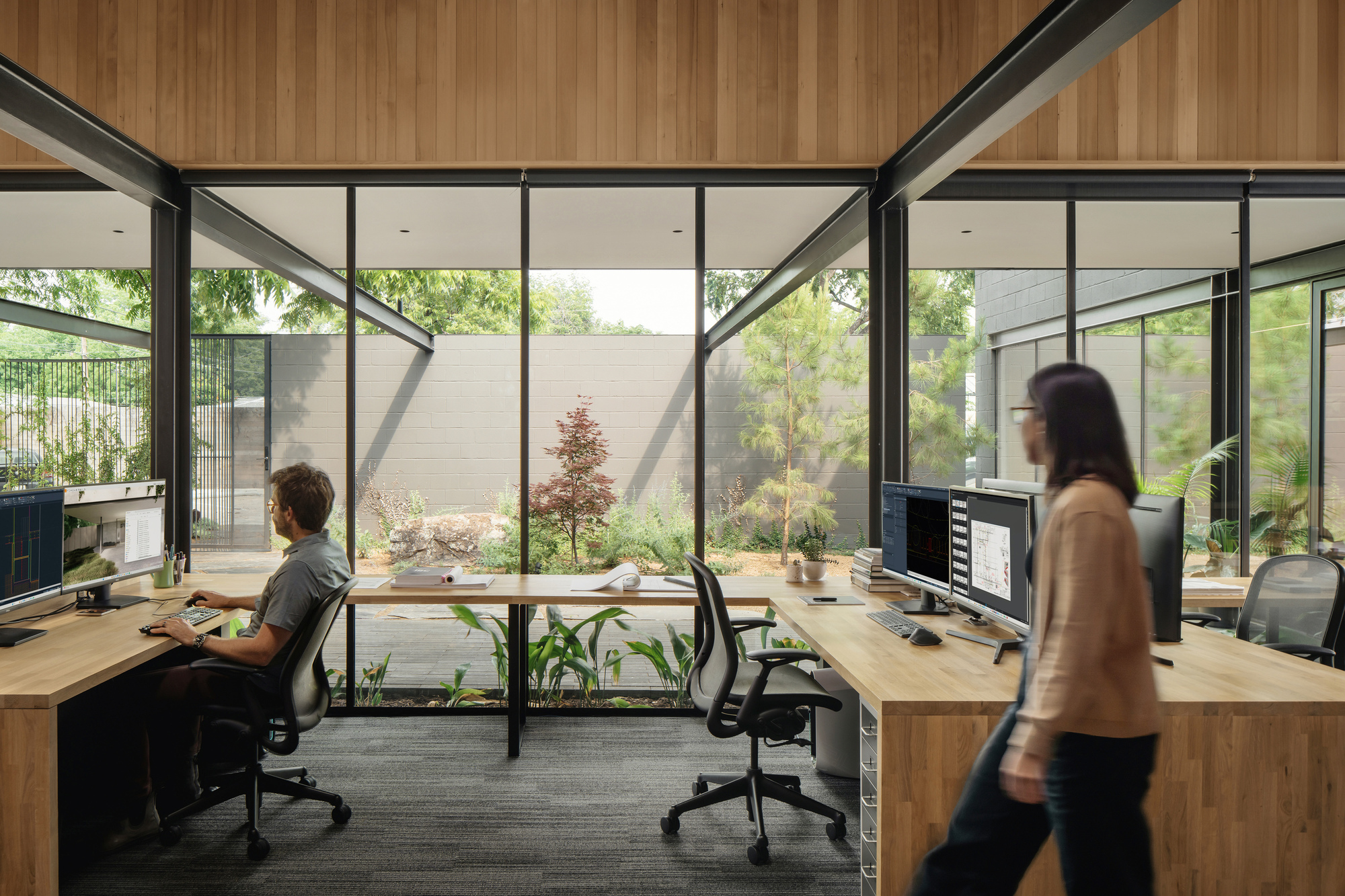 Open studio with timber desks and monitors facing a courtyard garden, one person seated and another walking past
