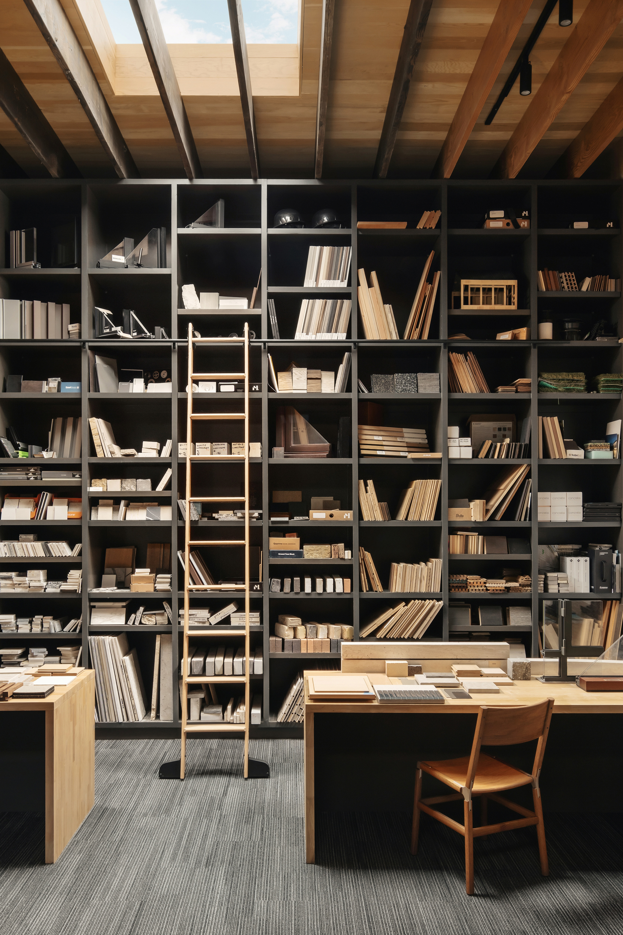 Floor-to-ceiling black shelving of material samples with a wooden rolling ladder beneath a skylight between exposed timber beams