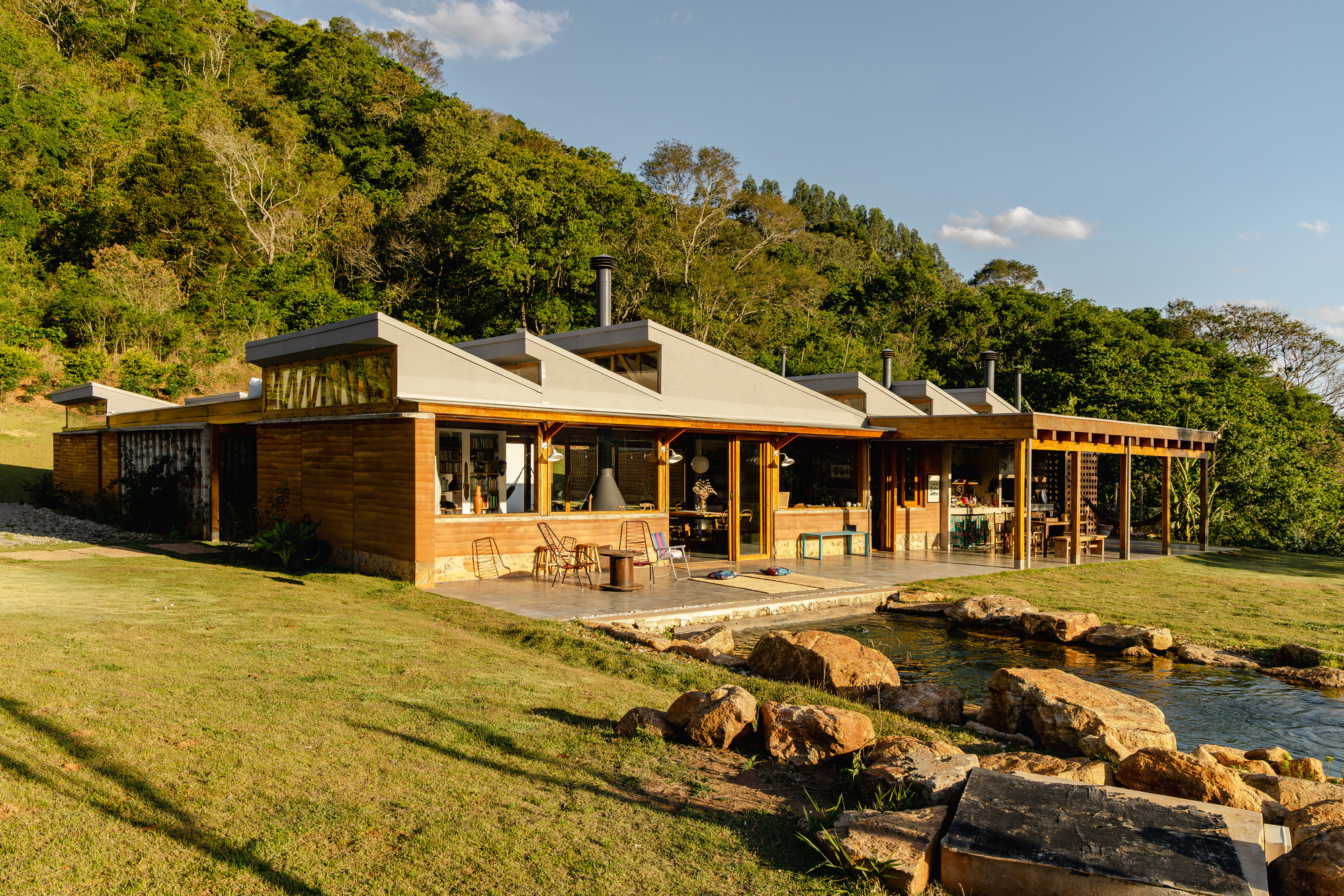 Low timber-clad house with angled roof monitors and a glazed living pavilion beside a rock-edged pond, lit by low golden sun