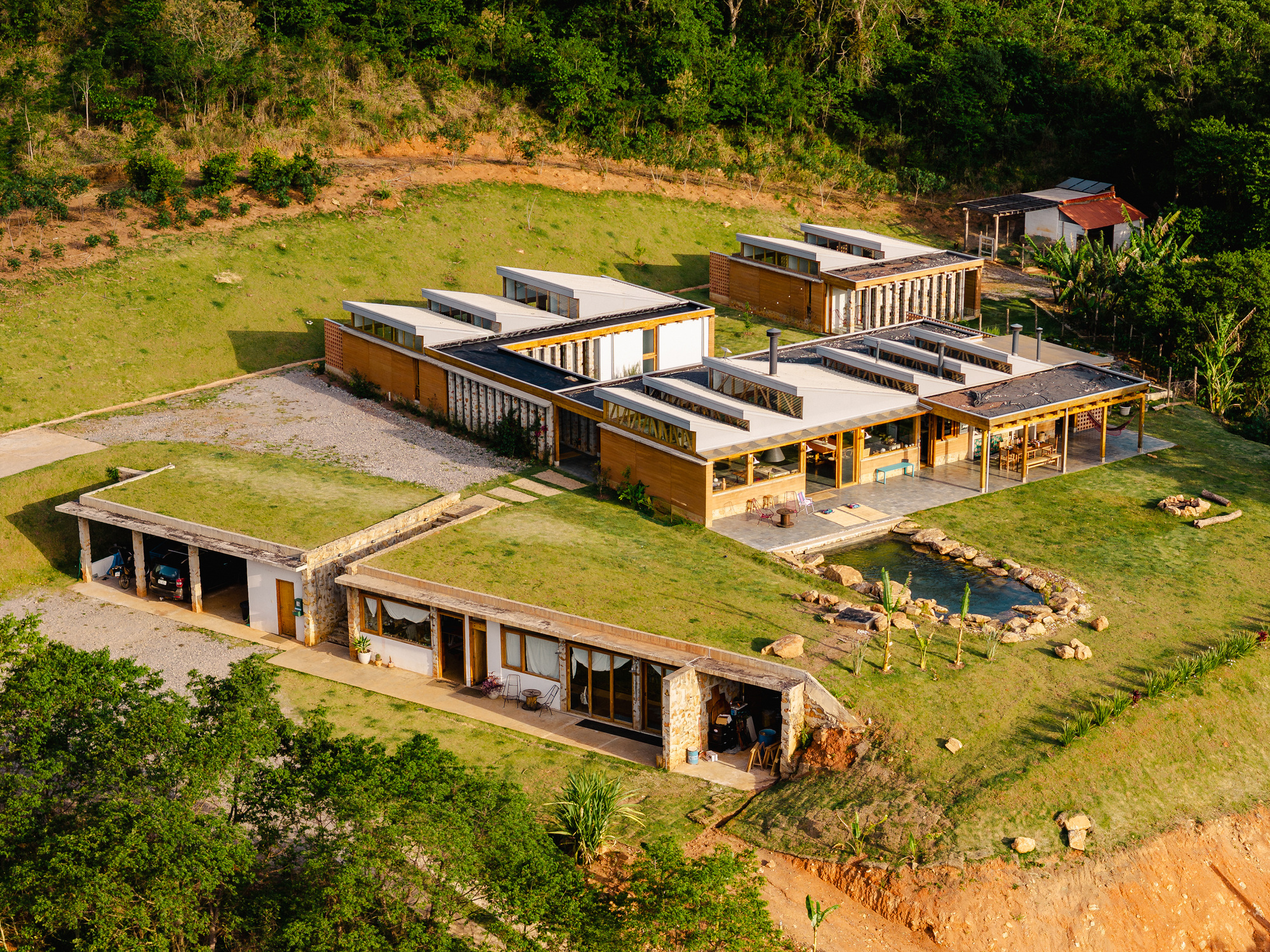 Aerial view of the long timber and stone house with sawtooth roofs and grass-roofed outbuildings on a green hillside
