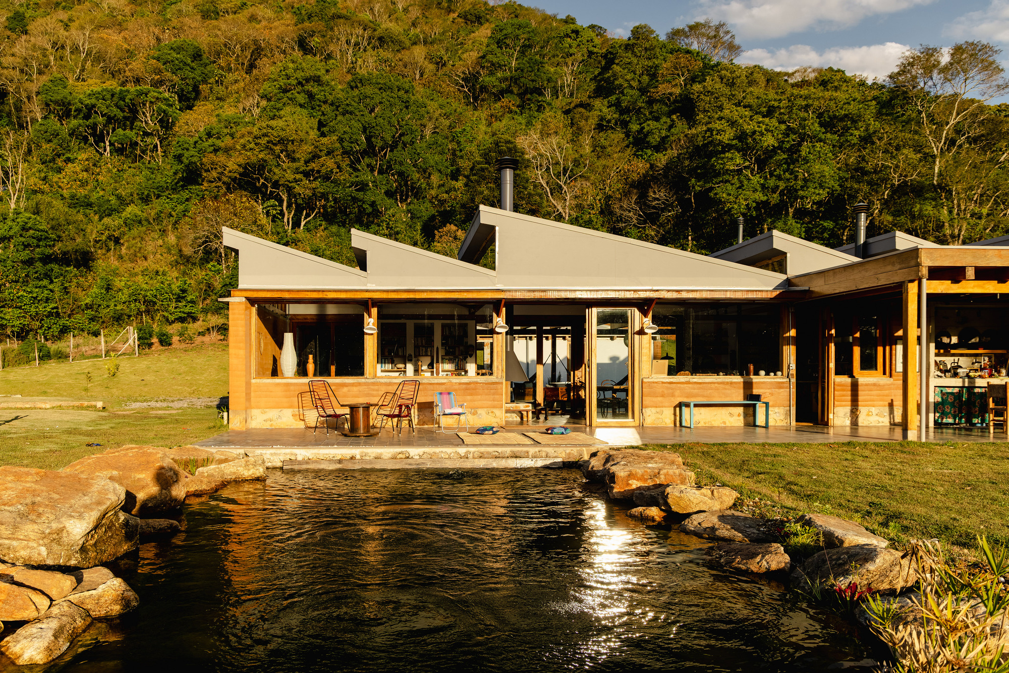 Glazed living pavilion with sloped roofs and a chimney flue reflected in a still pond ringed by boulders at sunset