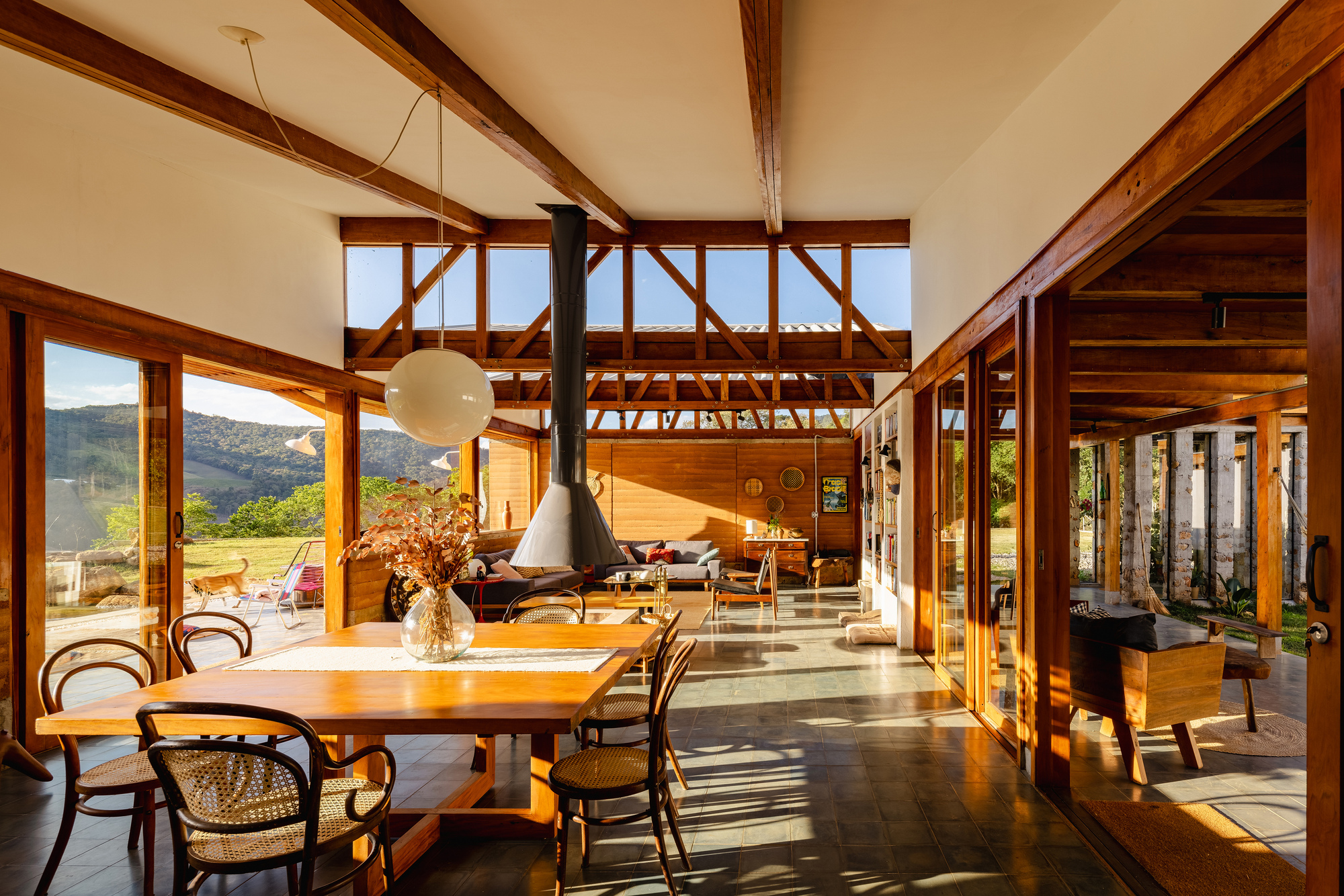 Open dining and living space with exposed timber trusses, a hanging black fireplace hood and sliding glass doors in afternoon sun