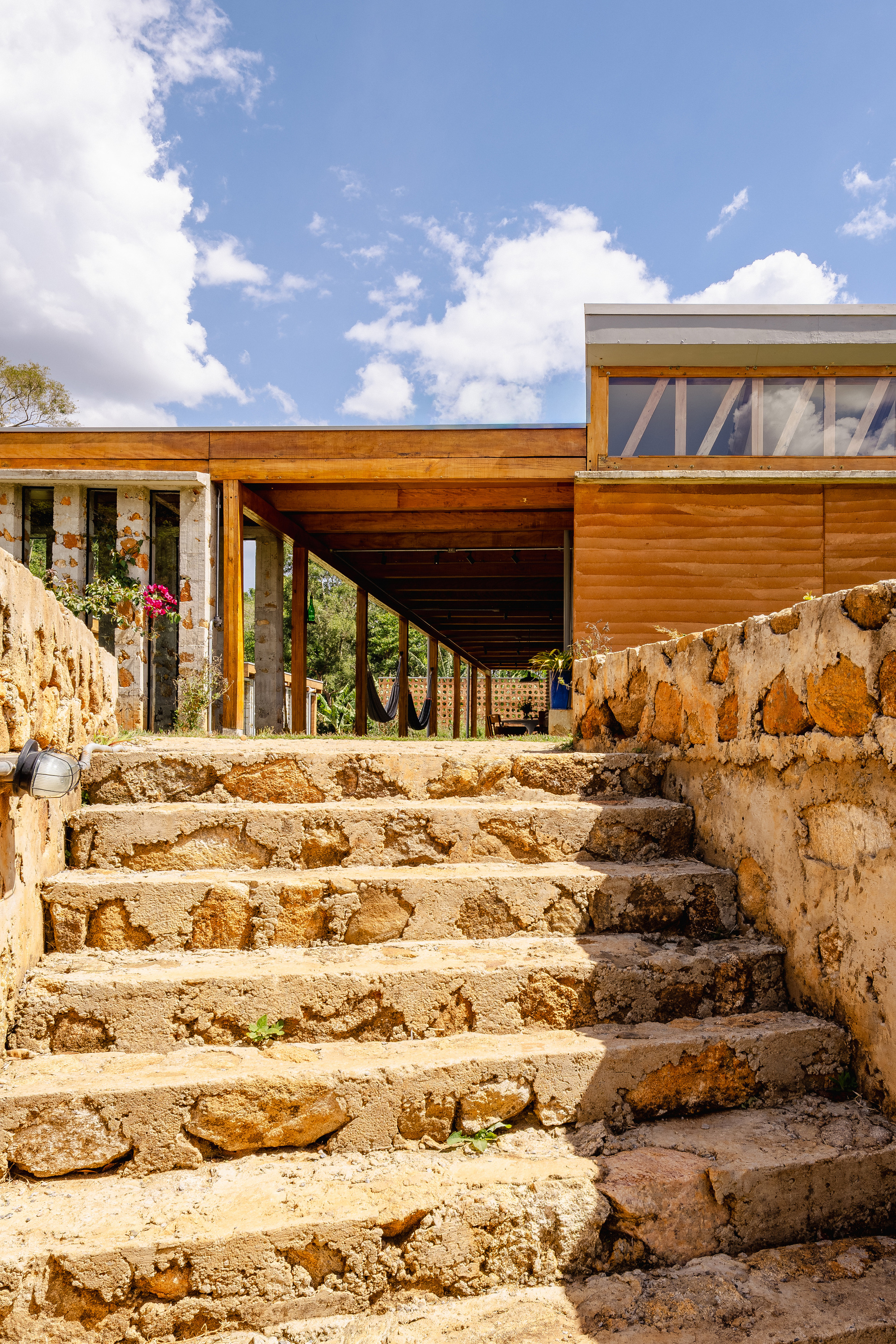Rough stone stairs flanked by rammed stone walls rising toward a timber-roofed veranda with a hammock under a cloudy blue sky
