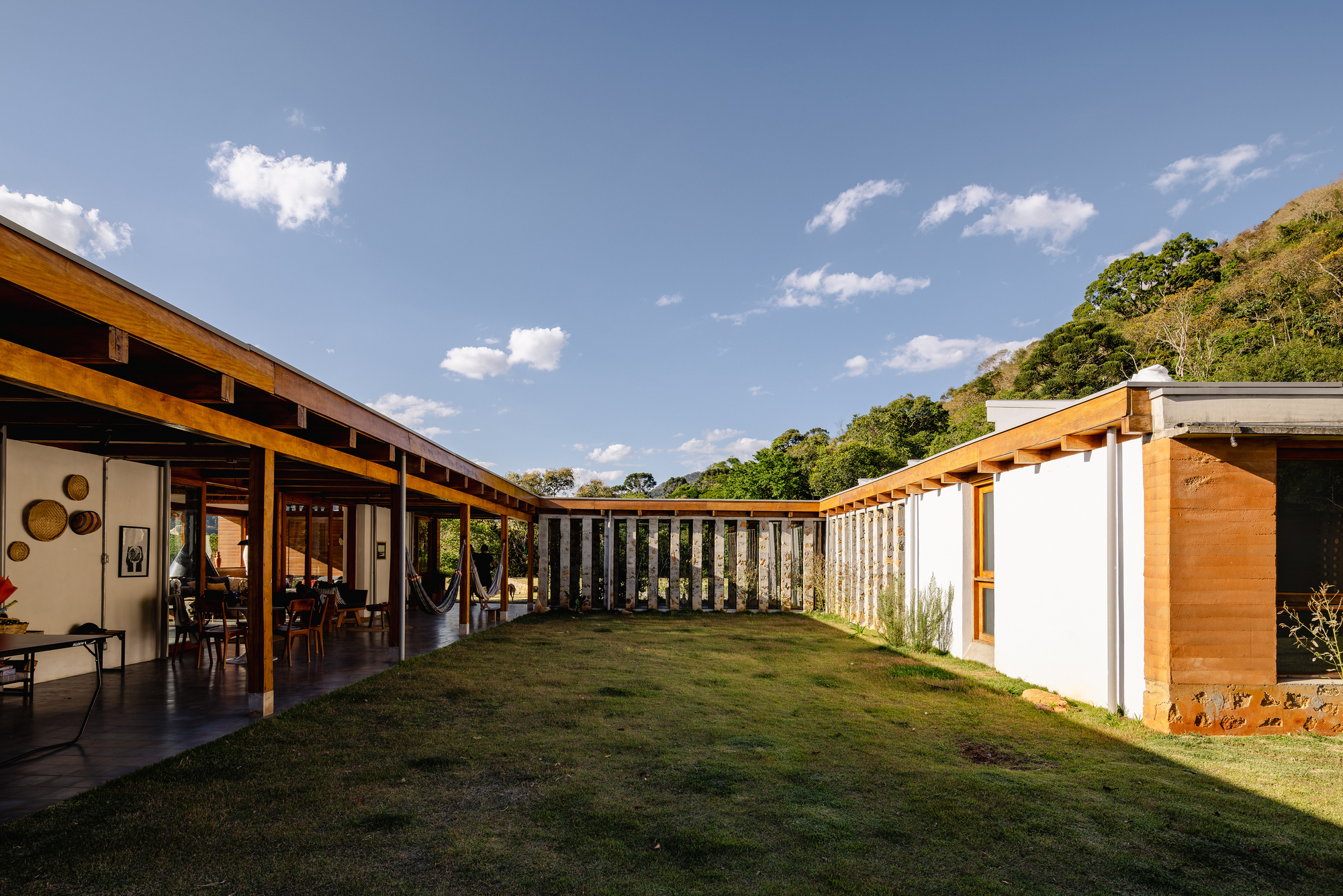 Grass courtyard framed by a timber-roofed veranda, rammed earth wall and a screen of slender stone piers under clear blue sky