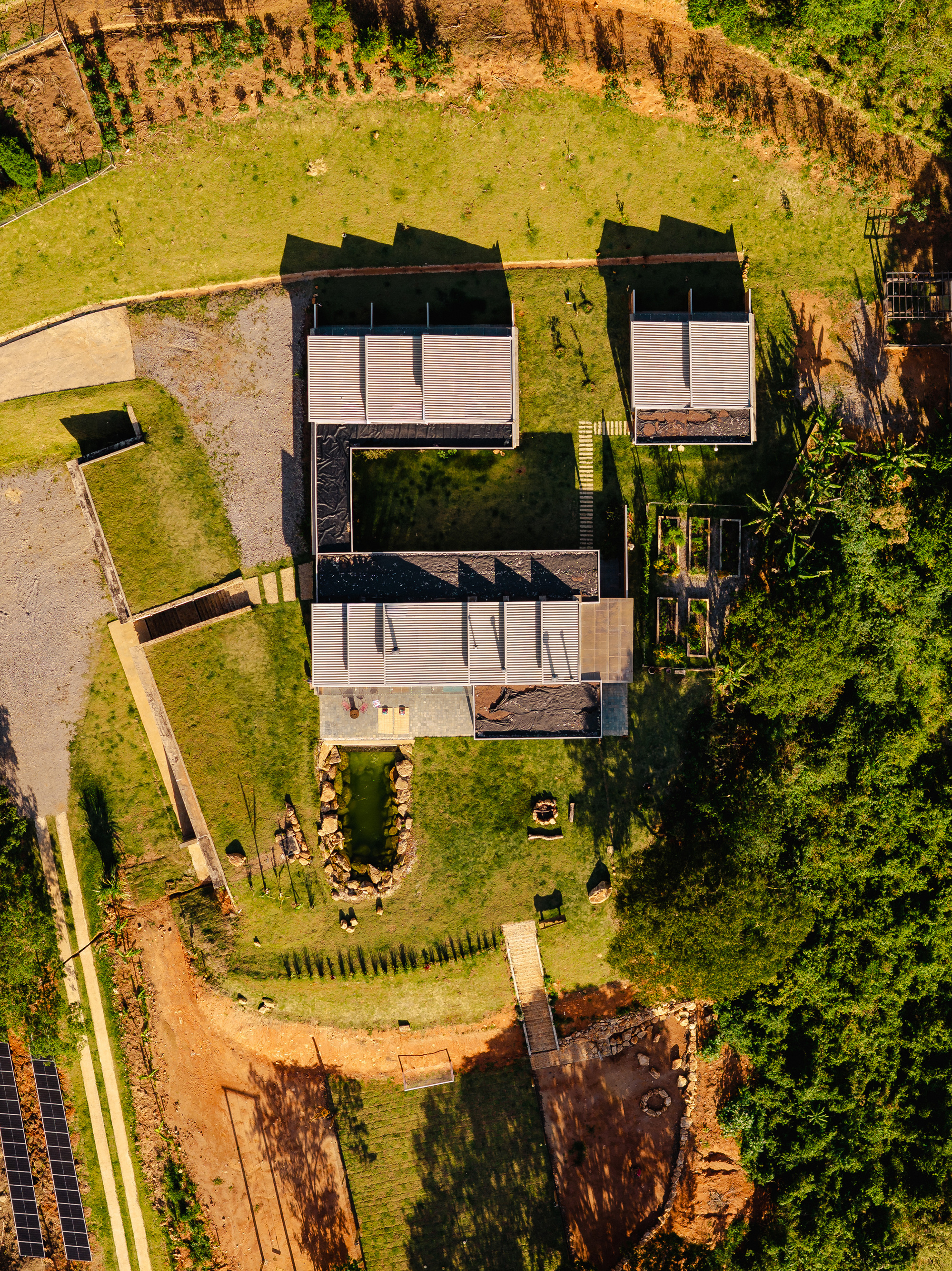Drone view of low pitched-roof pavilions around a courtyard, with a pond and long tree shadows on the hillside lawn