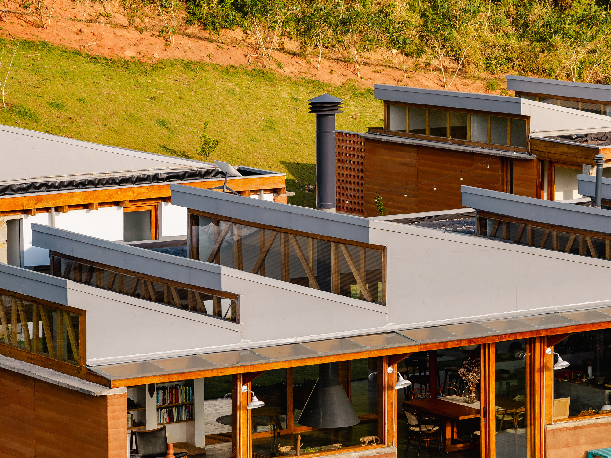 Close view of stepped sloping roofs with clerestory glazing, a black flue and a timber-framed dining room in low sun