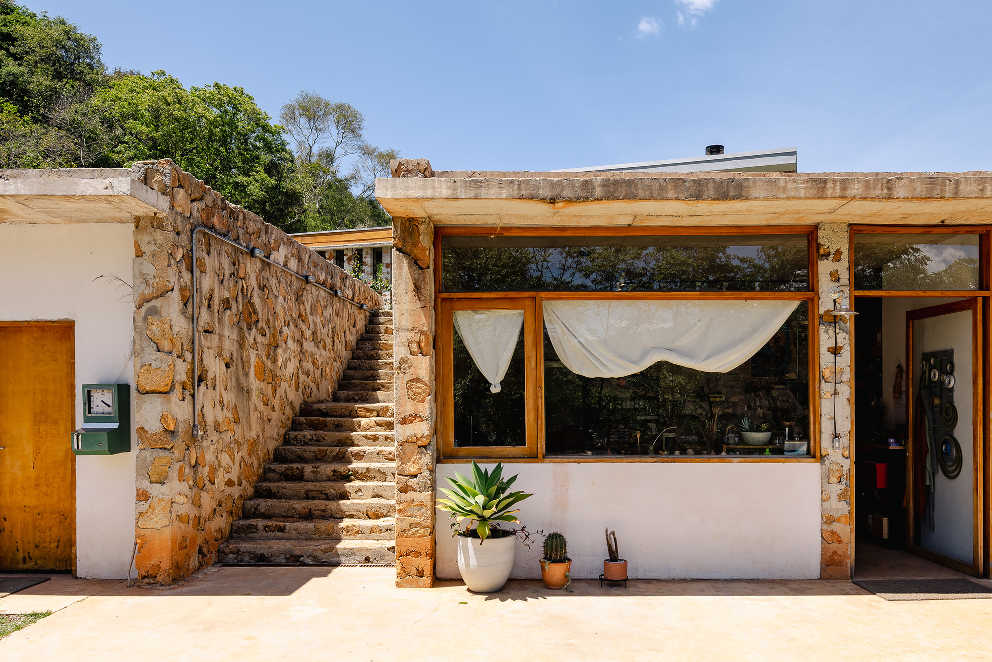 Stone-clad stair rising beside a timber-framed window wall with white curtains and potted agave in bright midday sun