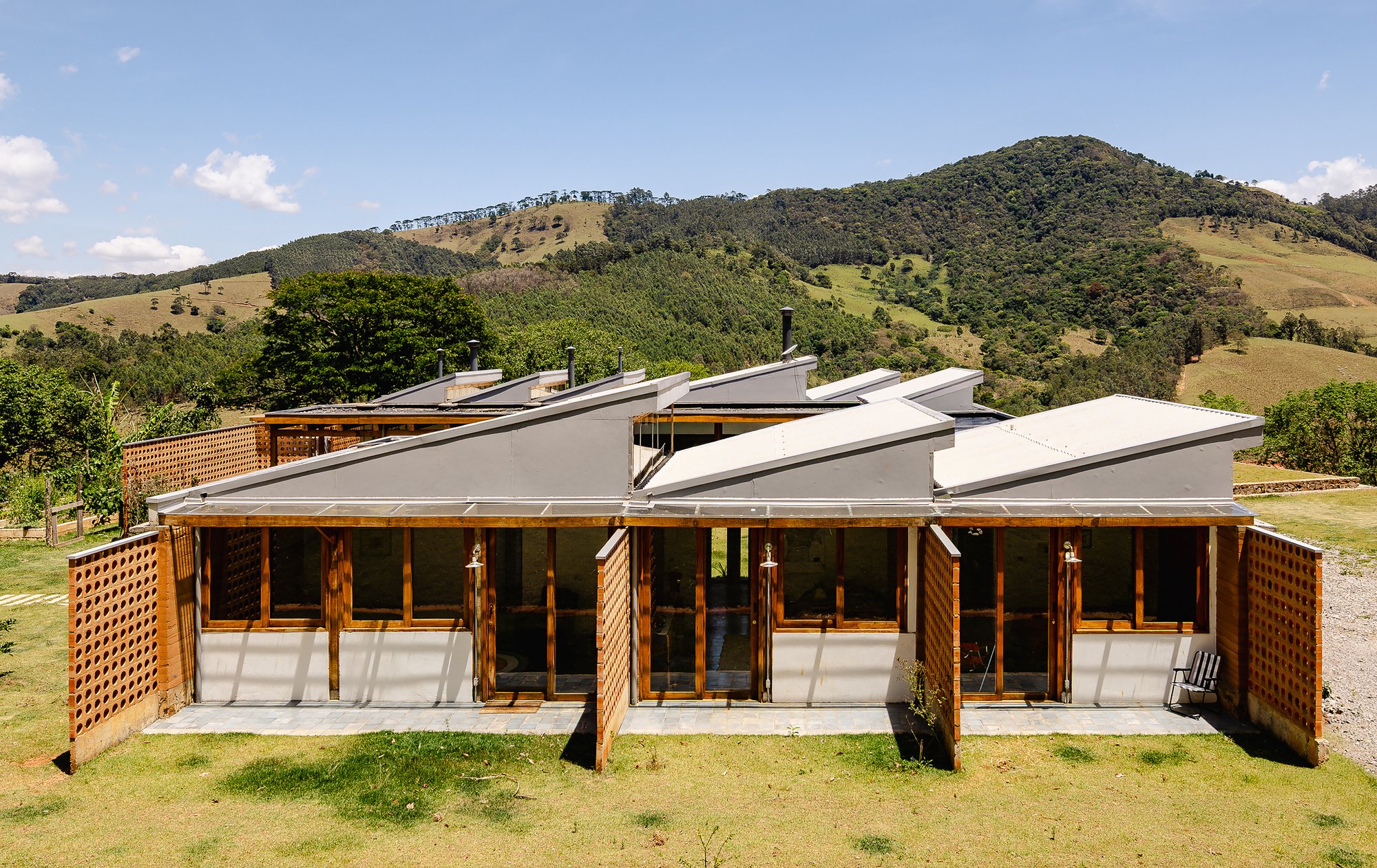 Front of a sawtooth-roofed pavilion with timber lattice screens and glazed doors, set against forested green hills
