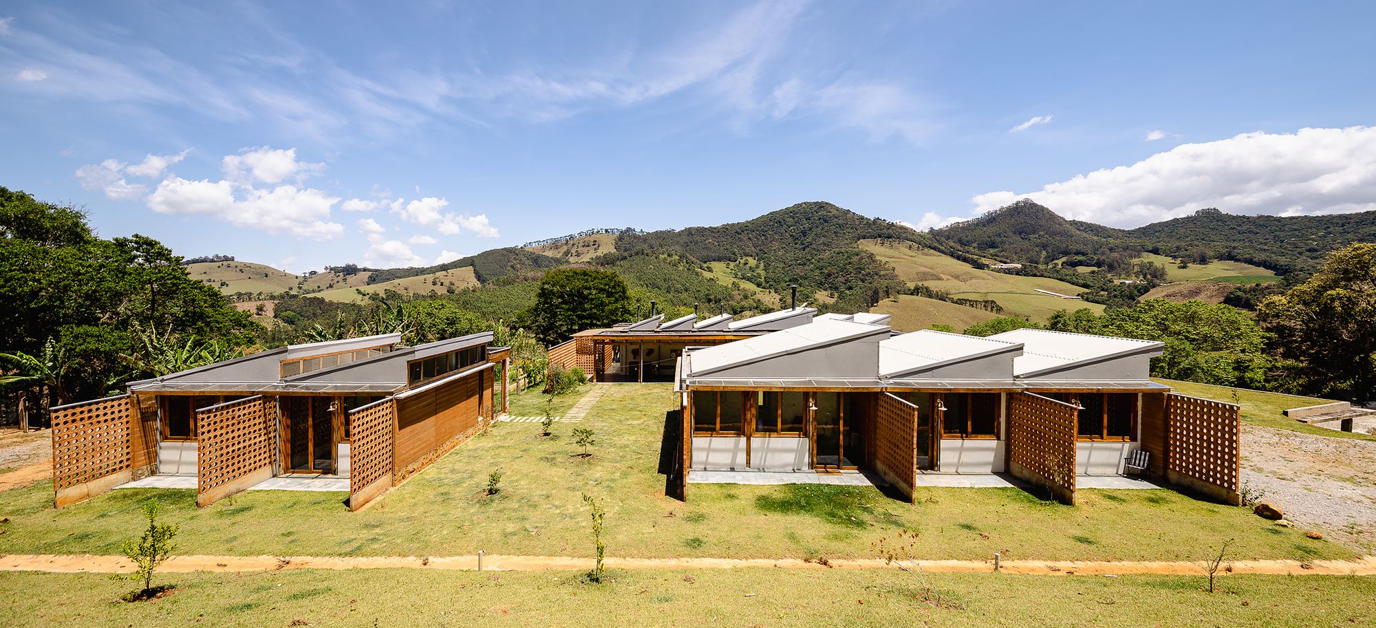 Wide view of two sawtooth-roofed pavilions with lattice shutters on a lawn, with rolling hills and wispy clouds behind