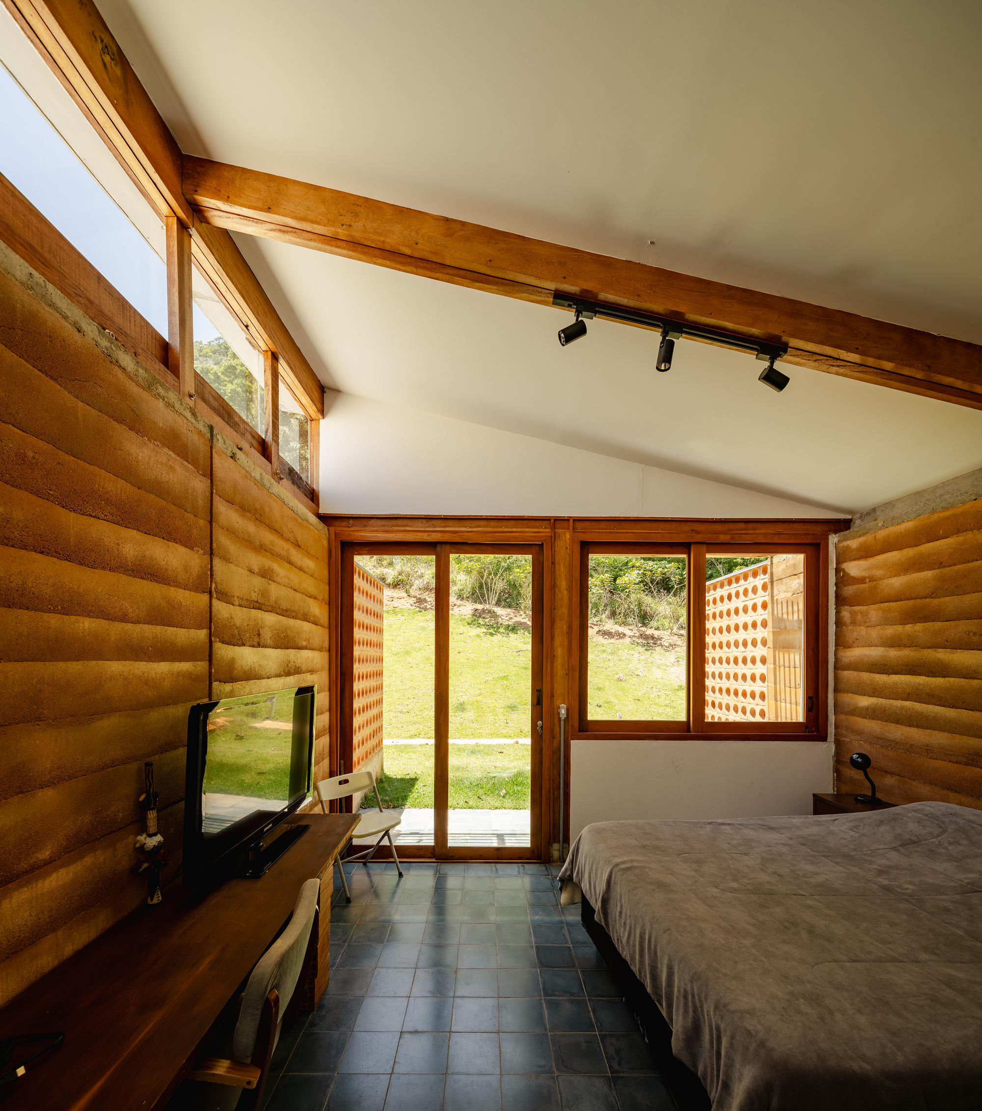 Bedroom with rammed earth walls, pitched timber beam ceiling and glazed doors opening to a sunlit lawn