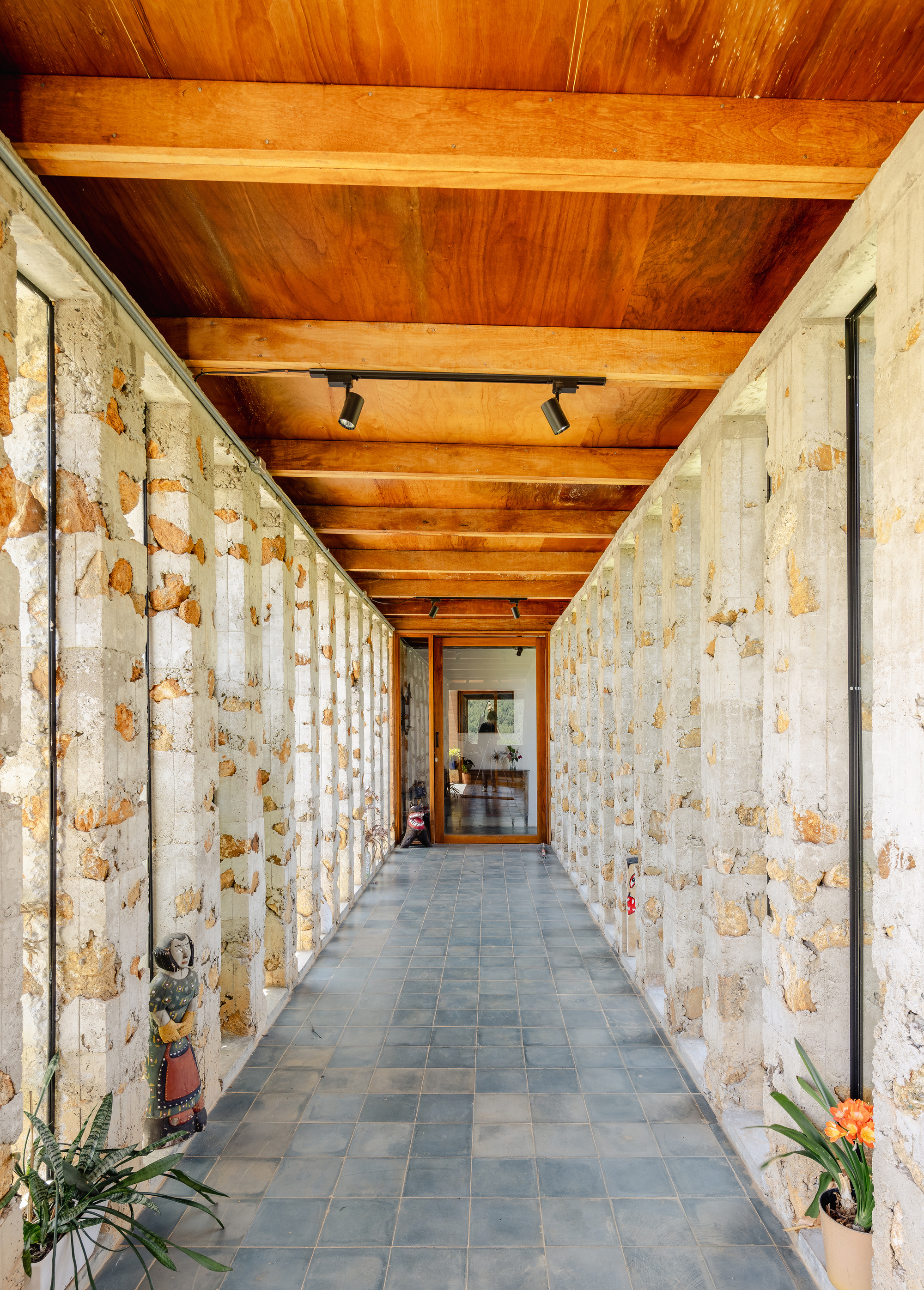 Long corridor lined with exposed stone piers and narrow glazing, slate floor leading to a glazed door and potted plants